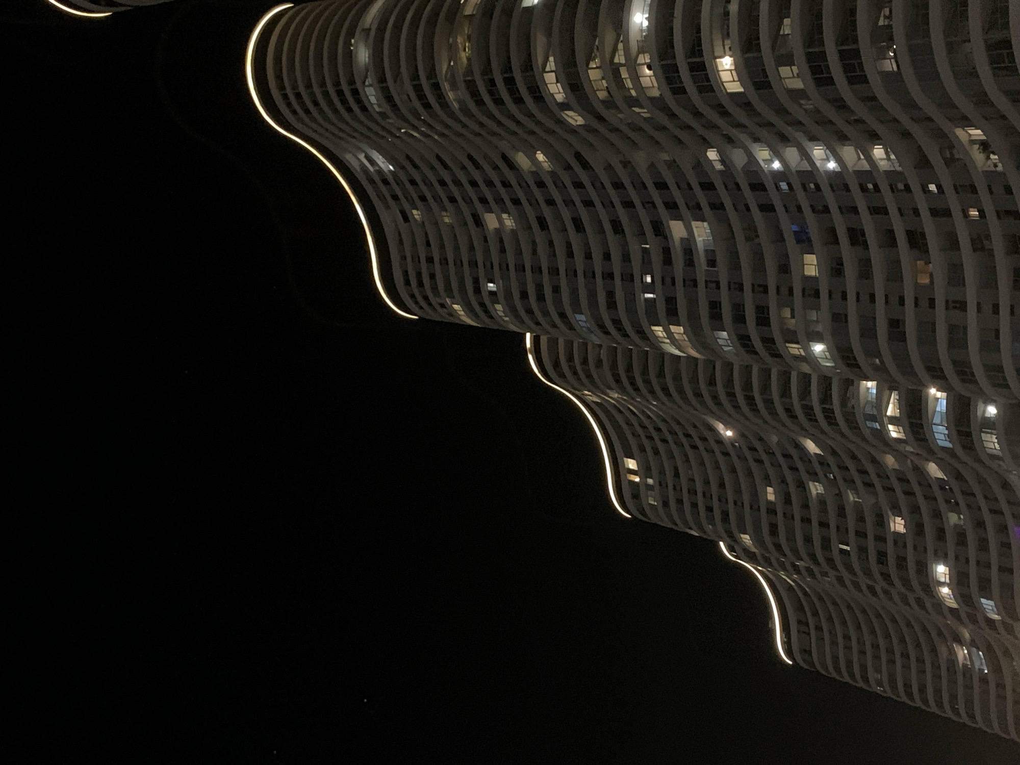 This is a nighttime, low-angle, full-frame photograph of a modern, wave-like apartment building in Tel-Aviv, Israel. The building features a distinctive undulating facade with balconies or protruding sections on each floor, creating a sinuous, organic shape. The building is illuminated by warm, yellow-orange LED lights that trace the curves along the top edges of its tiers. Numerous windows are visible, with some illuminated from within, suggesting occupancy. The sky is dark and appears mostly clear, with a faint hint of a distant light or star in the upper left quadrant. The overall impression is one of architectural dynamism and urban nighttime ambiance. The building's design is reminiscent of the iconic "City Gate" residential towers, located in the Neve Tzedek neighborhood of Tel Aviv.
