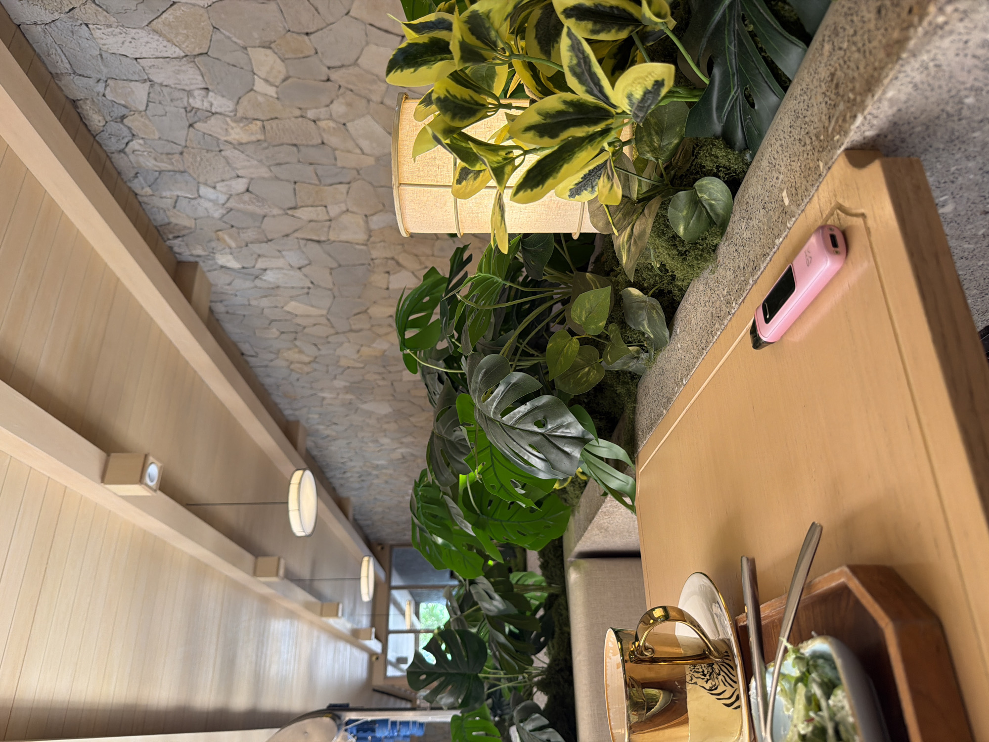 The image depicts the interior of a restaurant or cafe in North Kuta, Indonesia, featuring a modern design with natural elements. The ceiling has a textured, stone-like appearance. A wooden structure runs along the left side of the frame, punctuated by minimalist lighting fixtures. Plants cascade from a raised planter above a table. A lamp is placed amongst the plants. On the table, there is a pink electronic device, along with place settings including plates, silverware, and a cup. The scene suggests a stylish dining area focused on ambiance.