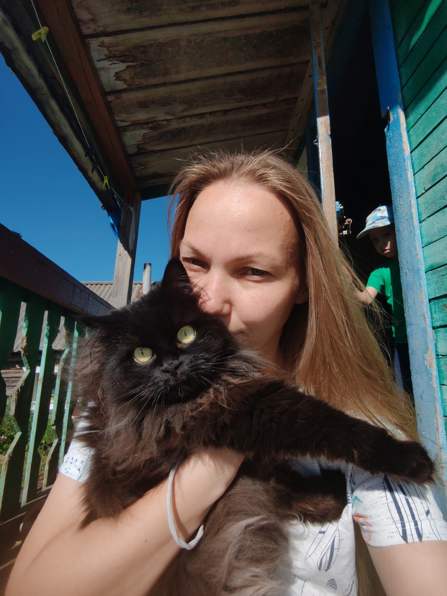 An adult female, appearing to be in her late 20s to early 30s with long blonde hair, is centered in the foreground, holding a fluffy black cat against her chest. Her face is partially obscured by the cat, with her right eye and the bridge of her nose visible, and she looks directly at the camera. She wears a white t-shirt with a repeating pattern of dark blue stylized feather or boat shapes. A white elastic bracelet is visible on her left wrist. The black cat has striking yellow-green eyes and very dense, long fur.
The scene is set on what appears to be a weathered wooden porch or balcony in Чухлэм, Russia. Overhead, a ceiling of aged wooden planks shows significant discoloration and wear. A thin white clothesline with two yellow clothespins is strung horizontally beneath this ceiling. To the viewer's left, a green-painted wooden railing with vertical slats runs parallel to the woman. Beyond this railing, a corrugated metal roof of another building is visible under a clear, bright blue sky. A white cylindrical chimney rises in the distance. To the viewer's right, a doorway framed by peeling blue-green painted wood reveals a dark interior. A young child, possibly male, wearing a green t-shirt and a light blue and white patterned baseball cap, is partially visible in this doorway, looking towards the camera. The overall lighting suggests a sunny day.