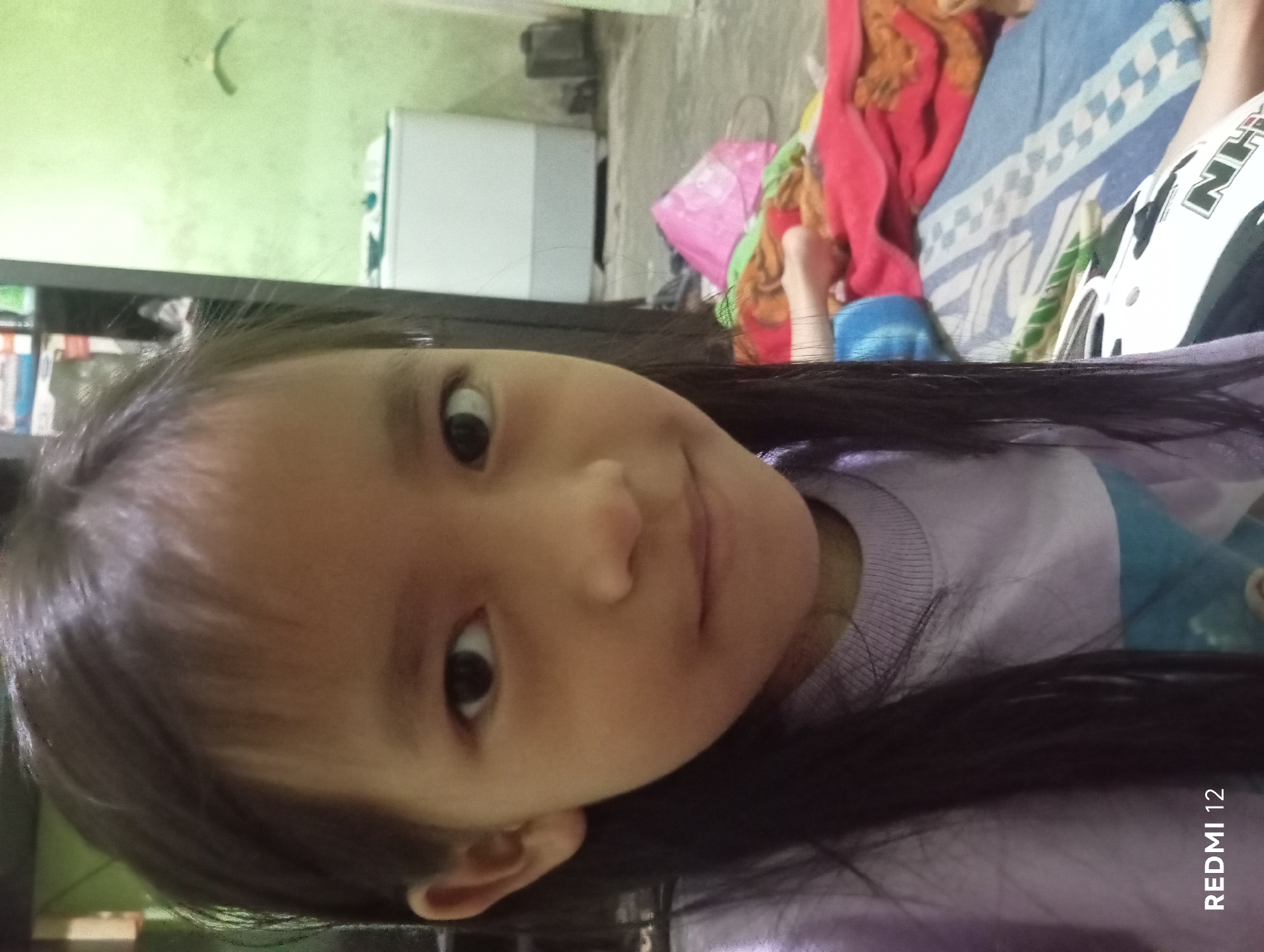 The image is a medium shot of a young East Asian girl in a purple sweatshirt, likely indoors. She is positioned in the foreground, with her face slightly turned to the left. Her dark hair is long and falls over her shoulders. In the background, a white washing machine is visible, along with a shelf unit containing various items. The floor appears to be tiled, and there are colorful fabrics and bags scattered around, suggesting a domestic environment. The lighting is somewhat dim, creating a slightly grainy effect. The "REDMI 12" watermark is present in the bottom left corner. Without additional information or context, it is impossible to determine the specific location within Pagar Alam, Indonesia.