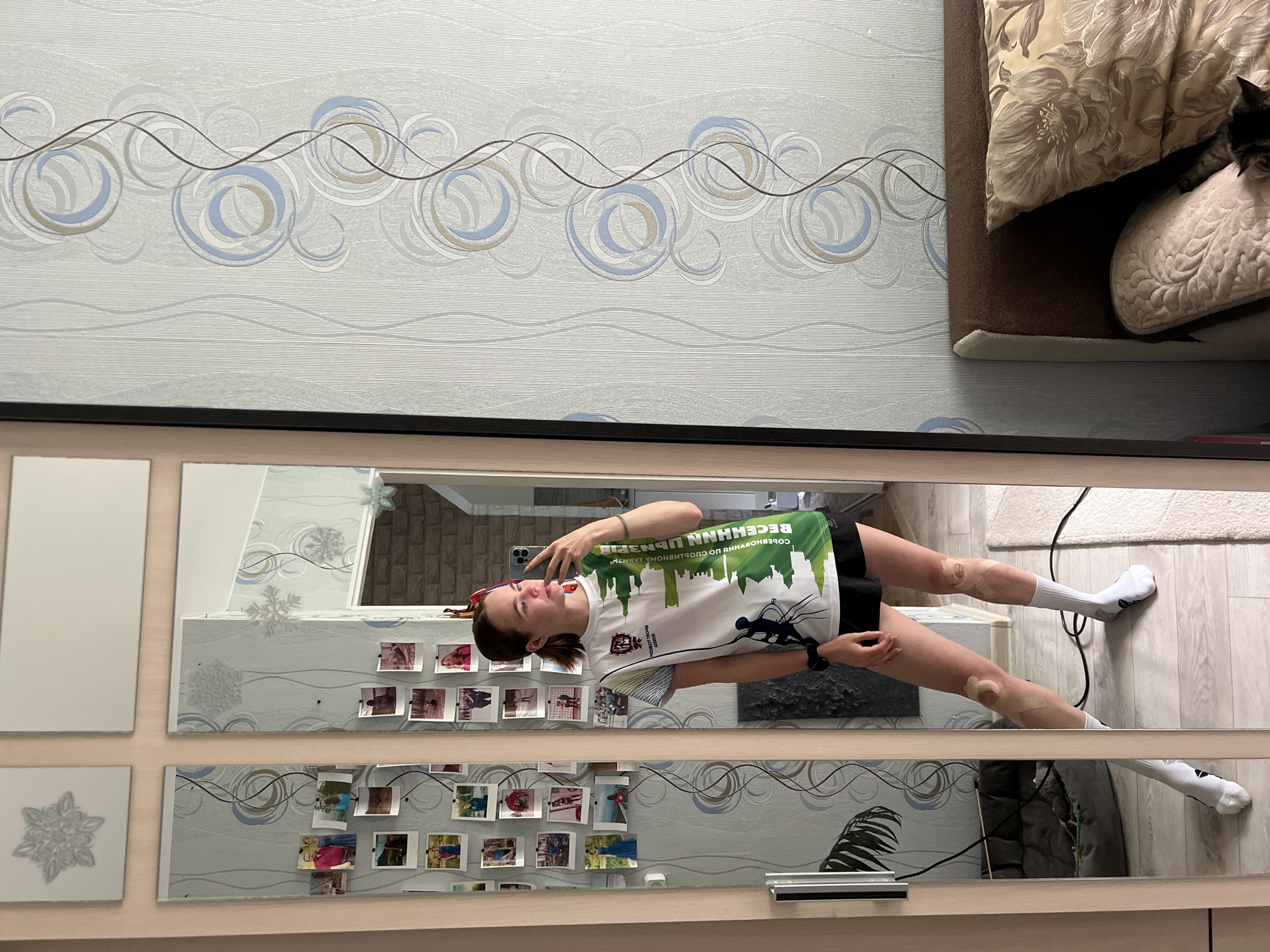 A young woman with brown hair pulled back in a ponytail is taking a mirror selfie in what appears to be a bedroom. She is wearing a white t-shirt with green and black graphics and black shorts. White socks are pulled up to her calves, and she has athletic tape on her knees. She is holding her phone up to take the picture and has a watch on her left wrist.
The mirror reflects a wall with light blue and white patterned wallpaper. Several small photos are attached to the mirror's frame on the left. The room also contains a bed with brown and patterned pillows, and a dark-colored cat can be seen partially on the bed. To the left of the woman, there is a dog bed.
The scene is set in an apartment in Chaikovsky, Russia, as indicated by the details in the wallpaper, furniture, and the presence of a personal living space.
