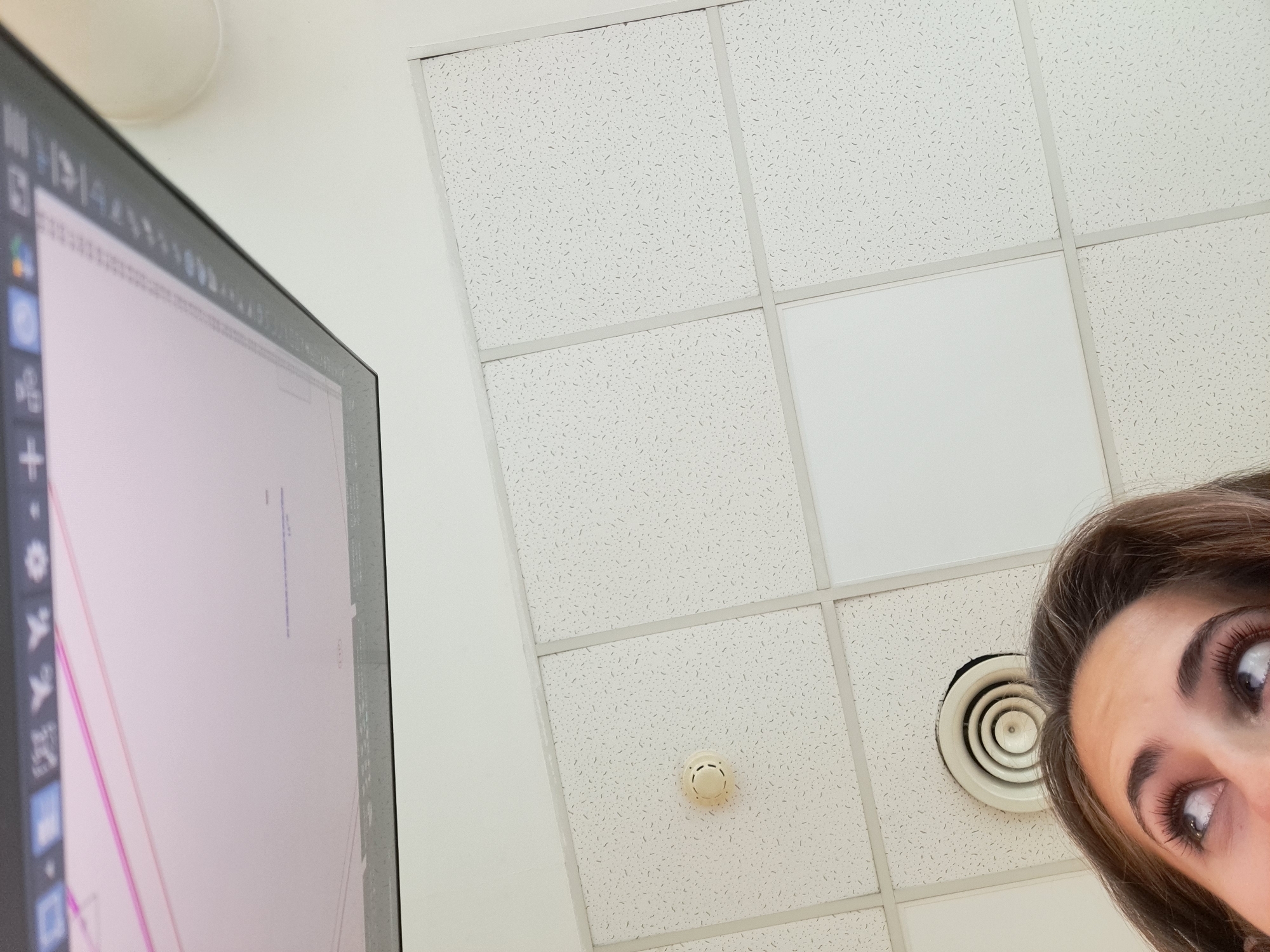 The image depicts a low-angle view within an office space. A computer monitor occupies the left side, displaying what appears to be a design or data visualization. Above and to the right, the ceiling is visible, composed of square acoustic tiles, one of which is plain white. An air vent and a smoke detector are mounted on the ceiling. In the lower right corner, a person's face is partially visible, with prominent eyes looking upwards. There is no information present to confirm the location as Sofia, Bulgaria.