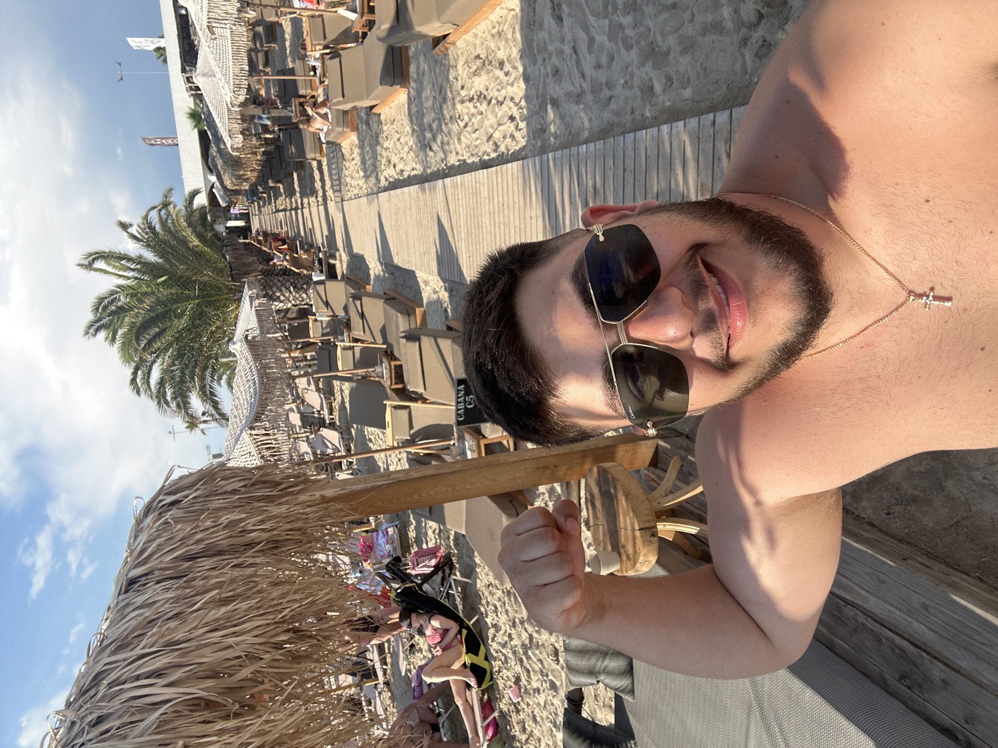 The image captures a man posing for a selfie on a sunny beach in Nea Kallikratia, Greece. The man, who is shirtless and wearing sunglasses and a gold cross necklace, flexes his bicep towards the camera. Behind him, a wooden walkway stretches along a beach lined with rows of lounge chairs and thatched umbrellas. Several other beachgoers are visible in the background, relaxing under umbrellas or walking along the pathway. Palm trees and a building with signage indicating "CABANA C5" are also present, suggesting a resort or beach club setting. The overall atmosphere is one of leisure and vacation.