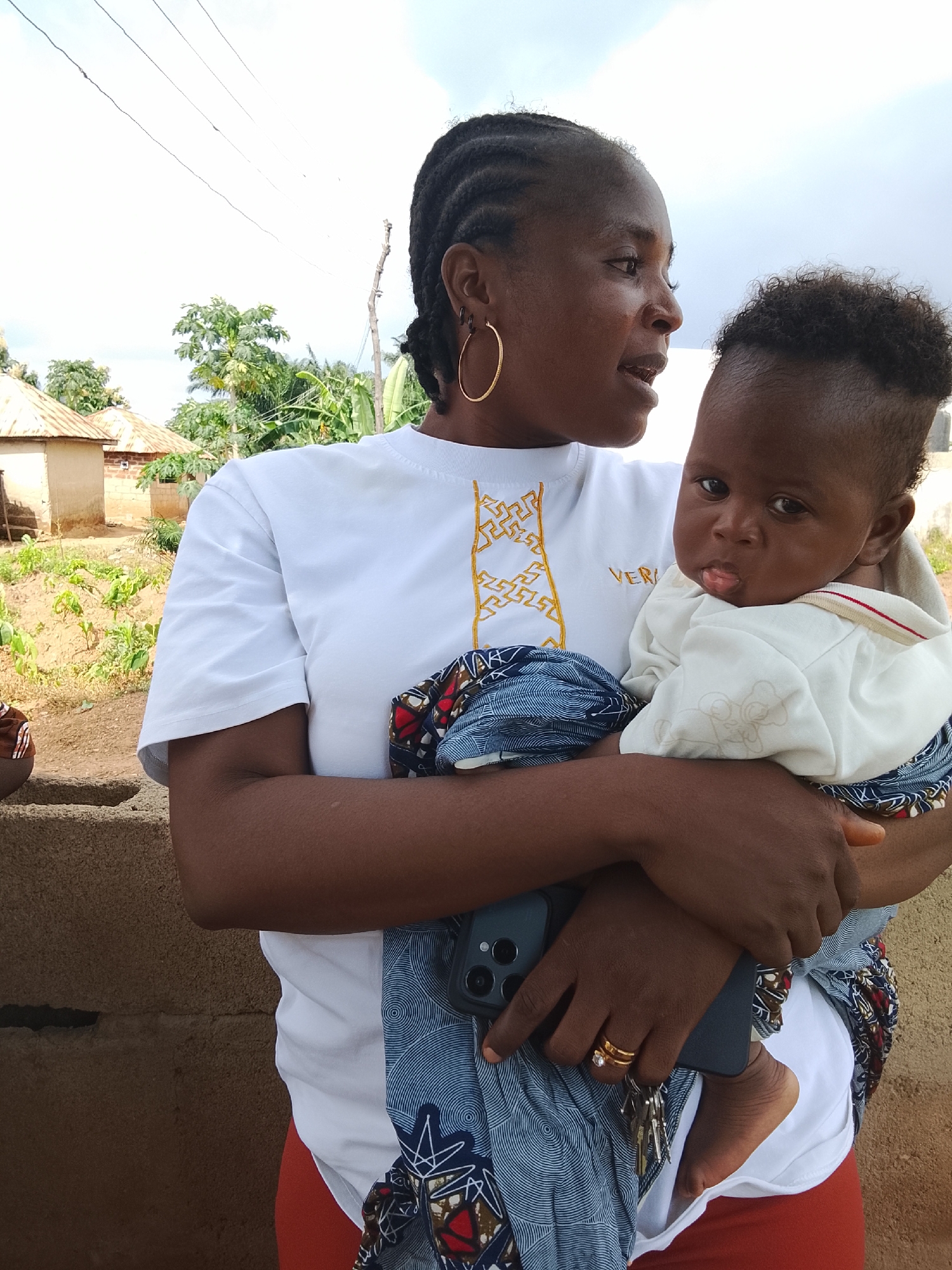 In Akwanga, Nigeria, a dark-skinned woman with braided hair is holding a baby. She wears a white t-shirt with gold trim and hoop earrings. The baby, also dark-skinned, wears a white outfit and is held close by the woman. Behind them, two small, thatched-roof buildings are visible, suggesting a rural or semi-rural setting. The ground is a mix of dirt and low-lying vegetation, with a stone wall in the foreground. The sky is overcast, with thin clouds and visible power lines overhead. The scene appears to be a casual, outdoor moment captured in a Nigerian village.