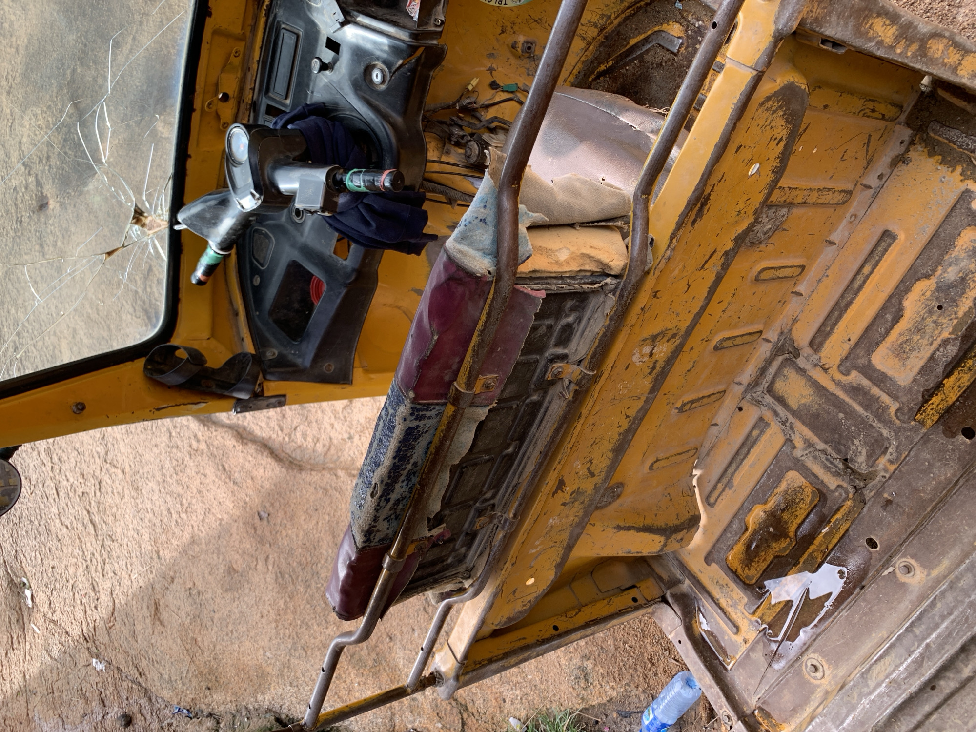 The image shows the interior of a yellow auto-rickshaw with its engine compartment and driver's seat visible. The windshield is cracked. The vehicle appears to be abandoned in a dusty, outdoor environment, possibly a roadside or junkyard, as indicated by the sandy ground and scattered debris. There is a blue plastic water bottle on the ground near the vehicle. The scene suggests neglect and disrepair, with rust and dirt accumulated on the vehicle's surfaces. The overall impression is one of abandonment or a vehicle awaiting disposal. The location in Jos, Nigeria is not directly ascertainable from the image itself but is provided as context.