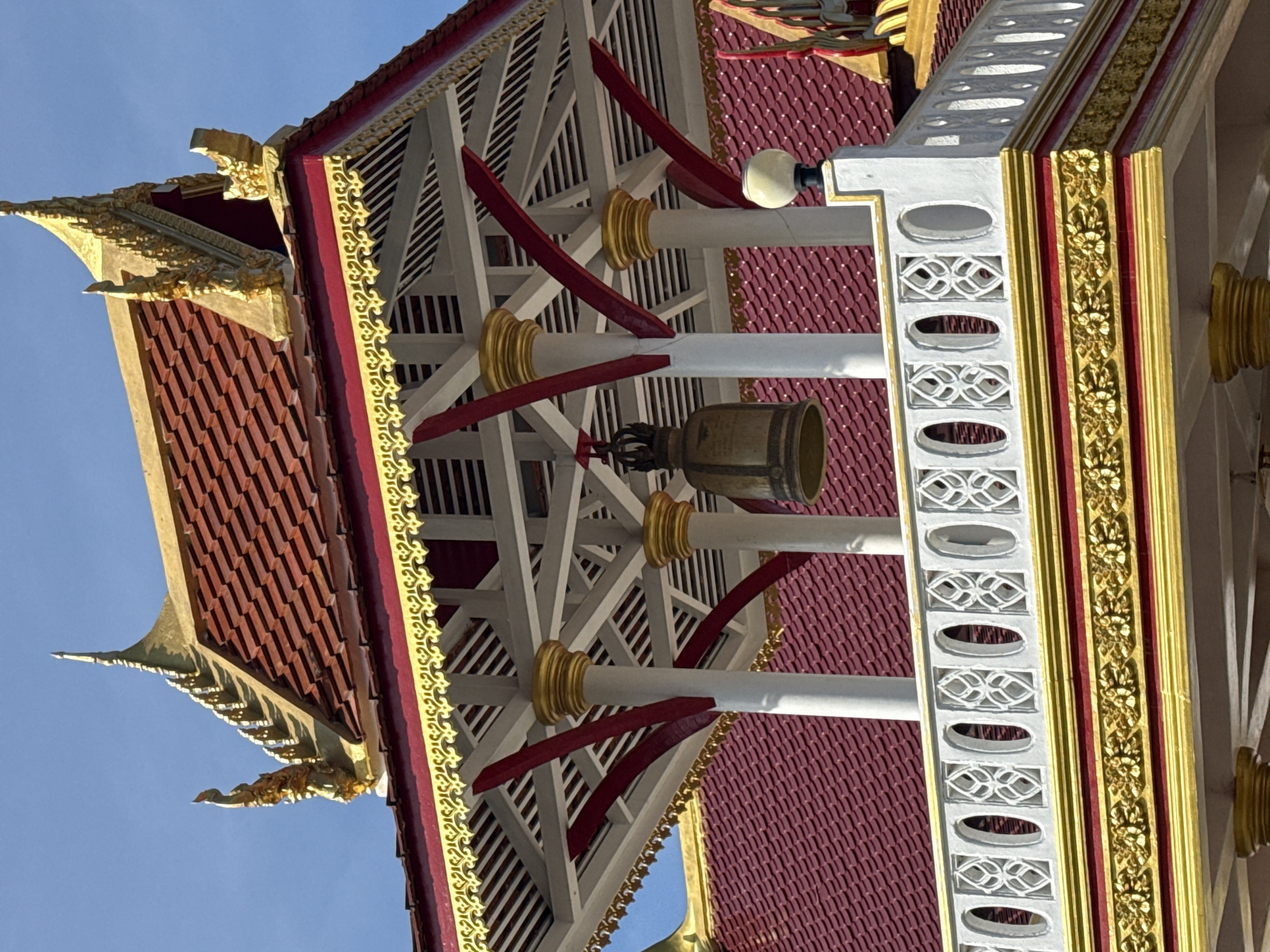 The image shows a close-up of the ornate roof and upper structure of a Buddhist temple, likely located in Baan Lamai, Thailand. The architectural style is characteristic of Thai temples, with multiple tiered roofs covered in red tiles and adorned with intricate gold detailing. The main focus is on a bell suspended within a white, open-sided pavilion supported by white columns topped with gold accents. The pavilion has a decorative white balustrade with an oval cutout pattern. Below the balustrade, there is a band of red trim decorated with a repeating pattern of gold floral motifs. The sky above is a clear, bright blue, suggesting a sunny day. There are no people or animals visible in the frame. The overall impression is one of grandeur and religious artistry.