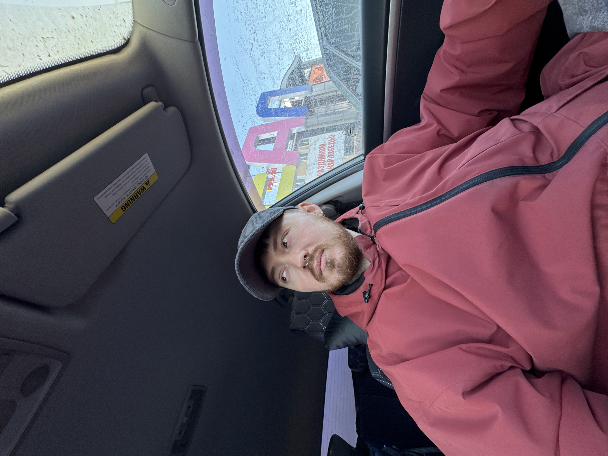 The image shows a man wearing a dark red jacket and a grey cap, seated inside a car. The man has a beard, a nose ring, and is looking towards the camera. Outside the car window, it appears to be raining. Reflected in the window is a building with a sign written in Russian: "РАЗДНИКОМ...НОЙ ПОБЕДЫ!" (Holiday...Victory!). A warning label on the car's sun visor is also visible. Based on the Russian text and the reflections outside the window, the location is most likely in the city of Chaikovsky, Russia.