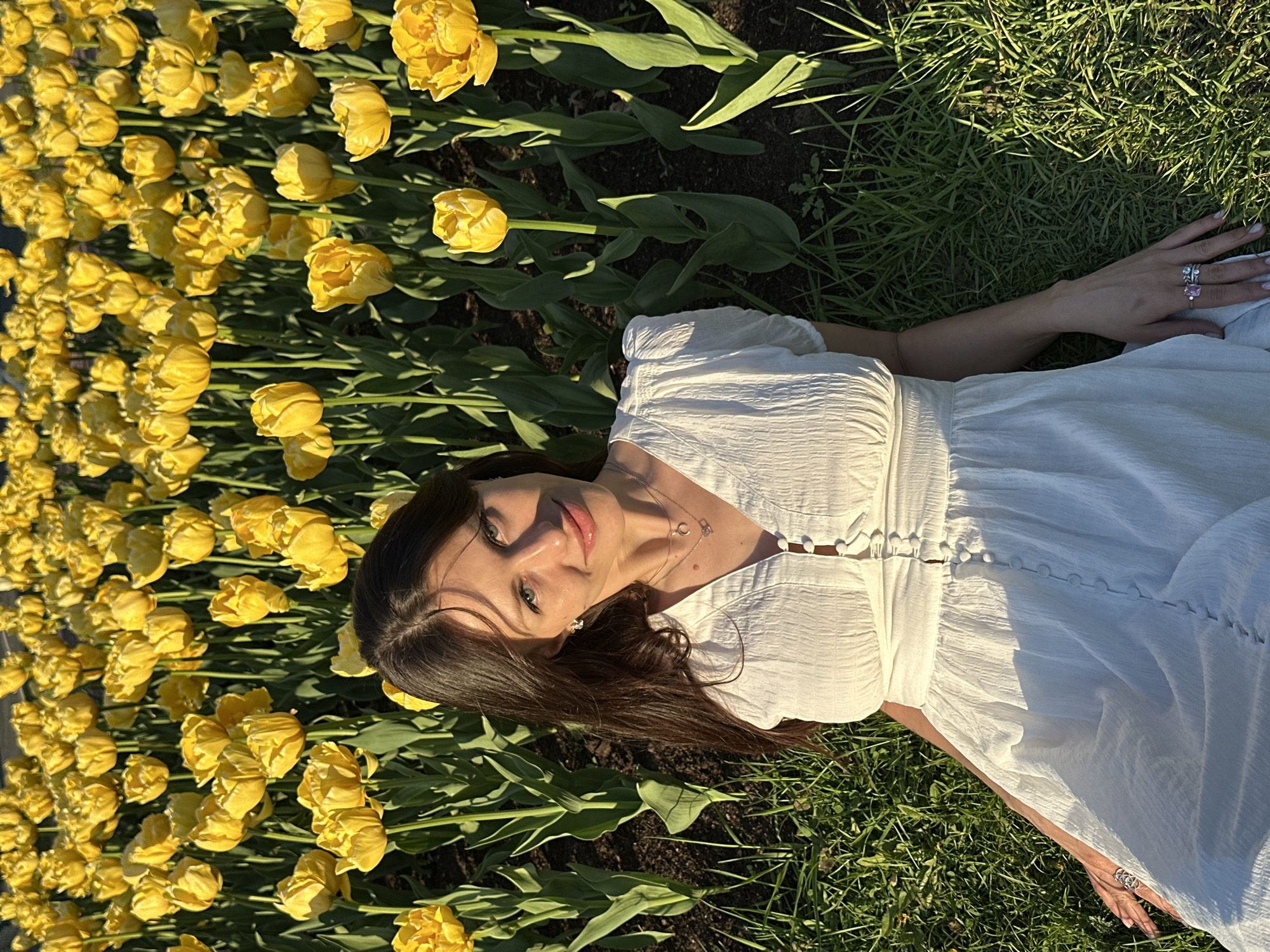 A woman in a white dress lies on the ground next to a bed of yellow tulips in Saint Petersburg, Russia. The ground is a mix of dirt and grass. The woman is facing the camera, and her dark hair is spread out on the ground. She is wearing a necklace. Her right hand is visible with several rings on her fingers. The image is taken from an elevated perspective. The lighting is bright and sunny, with shadows cast by the tulips.