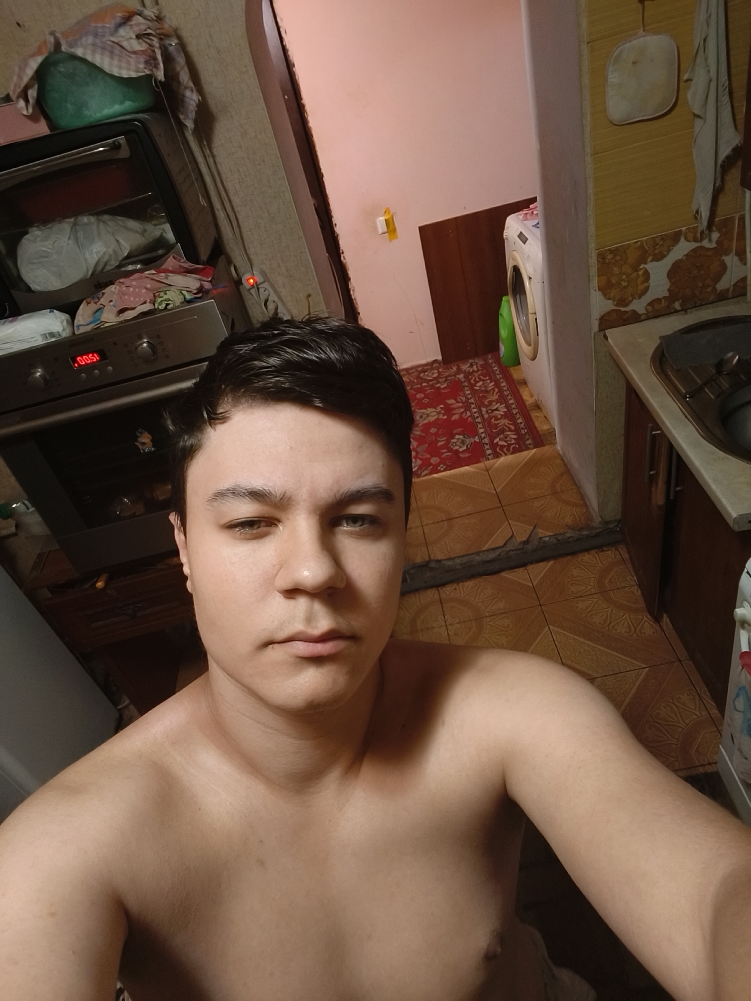 The image is a selfie taken in a domestic kitchen. The subject is a young male, shirtless, looking directly at the camera with a neutral expression. His left eye is slightly closed. In the background, an oven is visible, displaying a digital timer that reads "00.51". There is a red patterned rug on the tiled floor, and a washing machine is partially visible. The overall setting suggests a casual, home environment, possibly in the city of Chornobaivka, Ukraine, given the context of its use as a reference point. The scene appears to be indoors, with moderate lighting.
