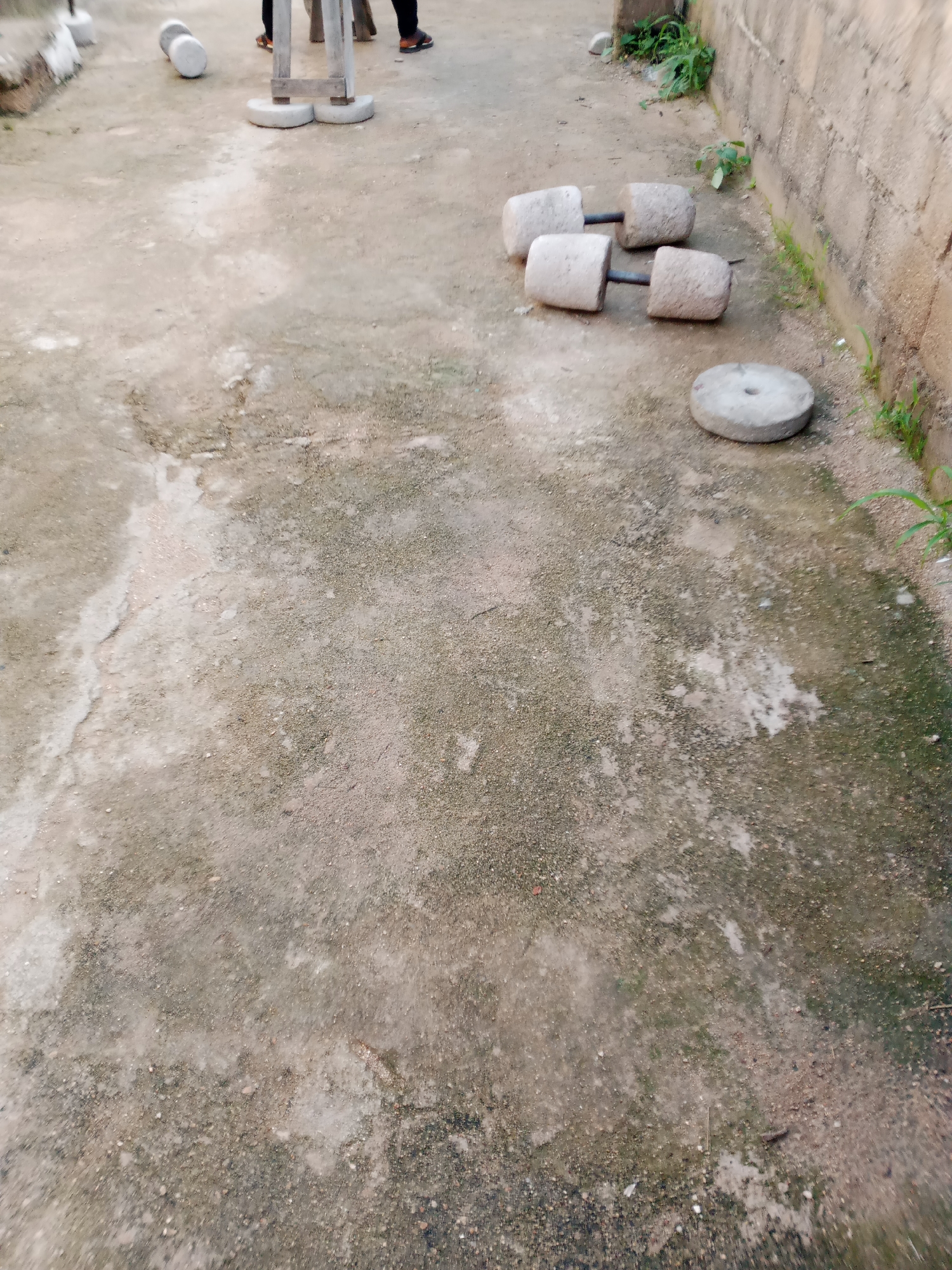 The image depicts an outdoor scene on a rough, concrete surface, likely in Jauro Yinu, Nigeria. In the foreground and middle ground, a pair of handmade dumbbells are present. These dumbbells are constructed from cylindrical concrete weights attached to metal bars. To the right of the dumbbells, a circular concrete weight with a central hole lies flat on the ground. The background shows a low, rough brick wall and some sparse vegetation. In the upper left of the image, a person's legs are visible, wearing dark pants and brown shoes, standing near some more exercise equipment. The overall impression is of an improvised outdoor gym or training area.