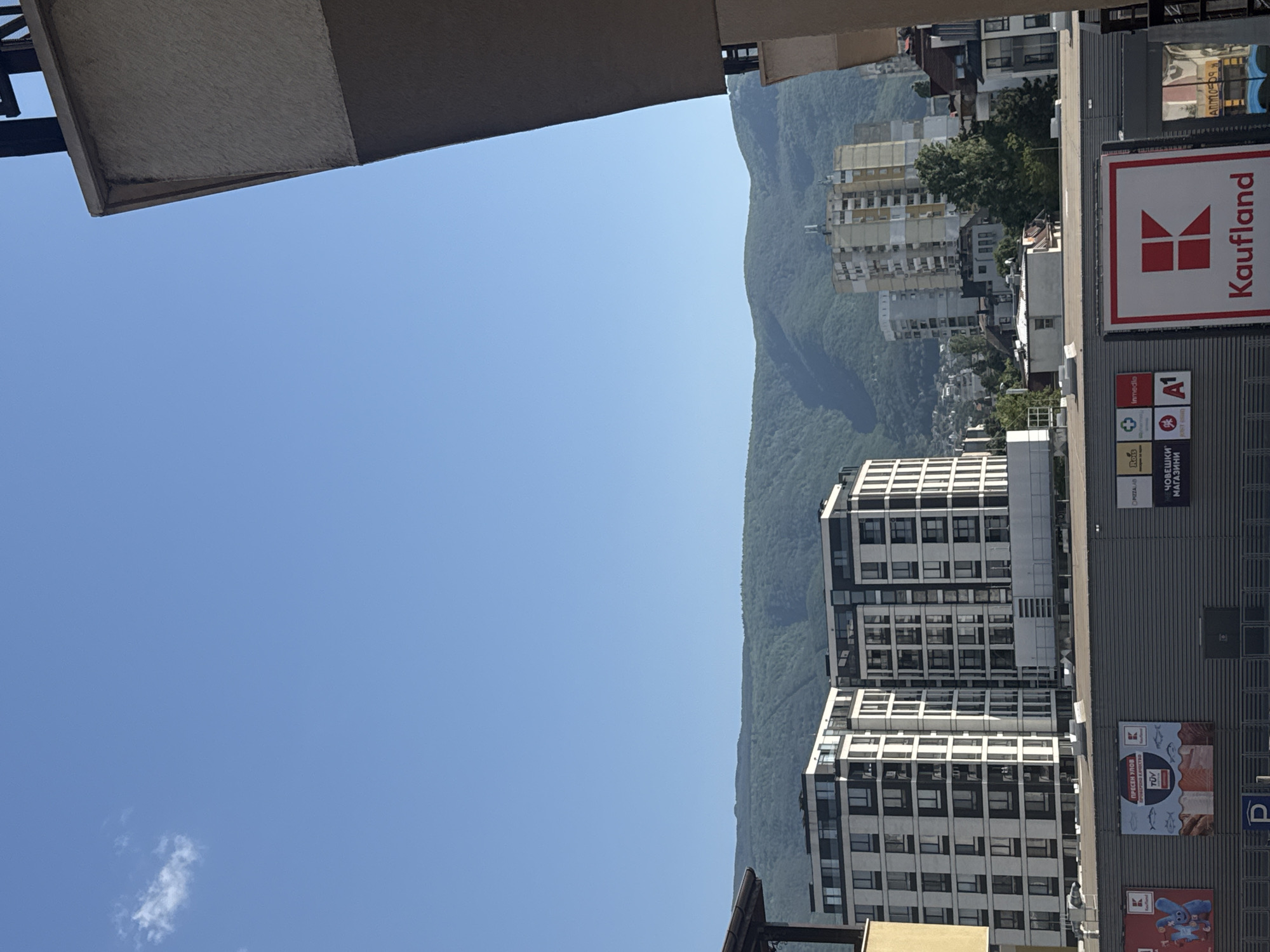 The image shows an urban landscape with a clear blue sky in Sofia, Bulgaria. The scene is dominated by the presence of modern buildings, including apartment complexes and commercial establishments. A densely forested mountain range forms the backdrop, providing a stark contrast to the urban environment. A prominent "Kaufland" supermarket building is visible, with its signage clearly displayed. Other smaller advertisements and logos are also present on the building's facade. The overall impression is of a vibrant city nestled against a natural mountainous landscape.