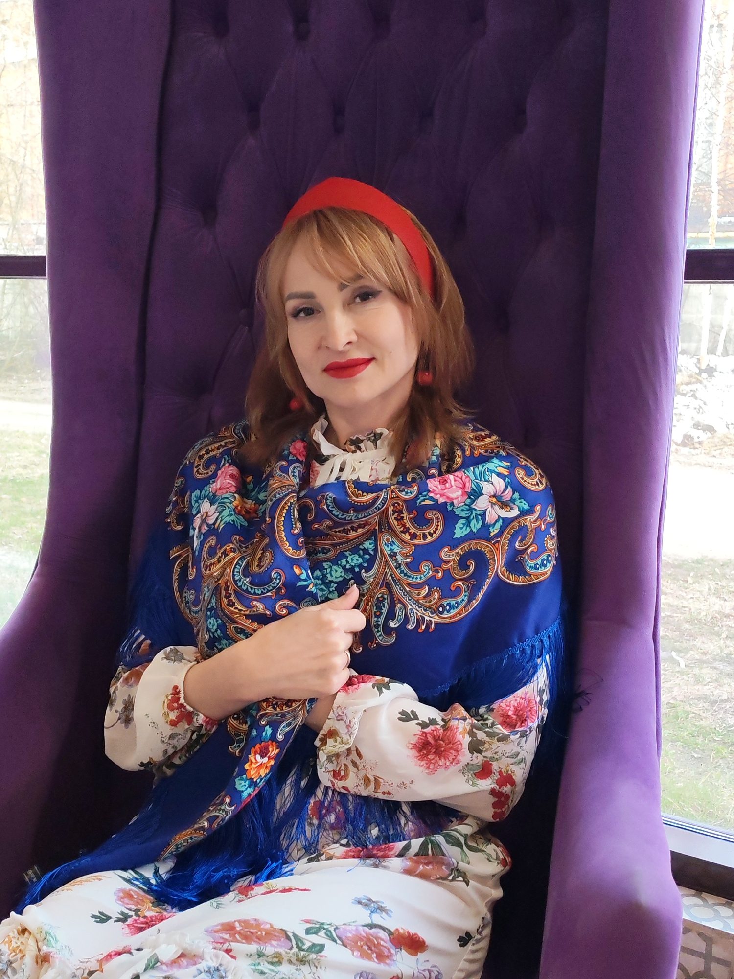 A woman with light brown hair and bangs is seated centrally in a plush, high-backed purple velvet armchair. She looks directly at the viewer with a pleasant, slight smile, her lips adorned with bright red lipstick that matches her red headband and small red drop earrings. She wears a white blouse or dress featuring a vibrant floral pattern with pink, orange, and green blooms. Over her shoulders is draped a striking blue shawl, richly decorated with intricate floral and paisley patterns in gold, pink, light blue, and green, complete with blue tassels along the edge. Her hands are gently clasped in front of her.
The armchair itself is ornate, upholstered in purple velvet with visible button tufting on the backrest. Large windows frame the woman on both sides, offering a view of an outdoor scene. The background outside appears to be natural, with bare trees or bushes visible, suggesting a daytime setting, possibly late autumn or winter. The light entering through the windows indicates it is daytime, though the weather appears to be overcast rather than brightly sunny. The style of the shawl is indicative of traditional Russian design, aligning with the provided location context of Chaikovsky, Russia. There is no visible text in the image.