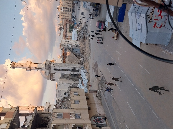 This image depicts a street scene in Khan Yunis, Palestinian Territory, under a partly cloudy, warm-toned sky indicative of late afternoon or early evening light. The central feature is a severely damaged white mosque with a distinct dome and a tall minaret, both exhibiting significant structural compromise, large holes, and missing sections. Surrounding the mosque are numerous multi-story buildings, also heavily damaged, with exposed interiors, shattered windows, and large piles of rubble at their bases.
The paved street in the midground has visible white road markings. Approximately 20-30 individuals are present on the street and sidewalks. In the foreground-middle, one person stands with arms outstretched, while others are seen walking individually or in small groups. A group of 8-10 people is gathered near a parked red car on the right side of the street. Another individual walks alone in the immediate foreground.
On the right side of the street in the foreground, a white tent with Arabic writing, including "نقطة سحن" and "حراج," is partially visible, along with a small wooden table. Overhead, dark utility lines or cables traverse the frame. The overall environment presents an urban landscape marked by extensive destruction and ongoing human activity amidst the debris.