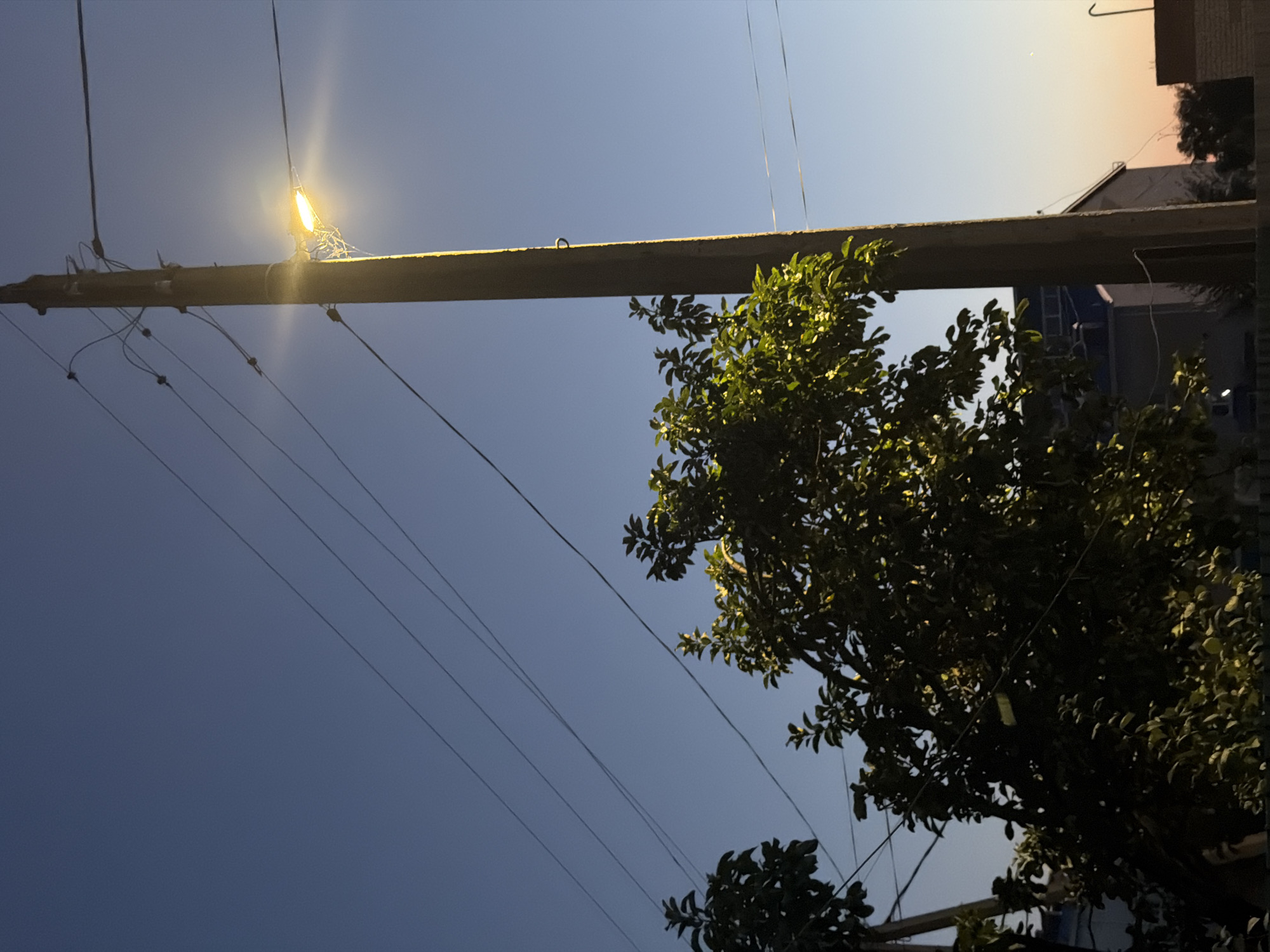 A vertical, weathered, wooden utility pole stands prominently in the center-right of the frame, illuminated by a bright, yellow streetlamp attached to its side. The lamp is casting a glow that creates a halo effect in the twilight sky. Multiple dark electrical wires extend from the pole and crisscross the scene, disappearing into the dusky blue sky. On the left side of the image, a dense, leafy green tree partially obscures the view of a low-rise building and a fence. The tree is lit from behind by the streetlamp, causing some leaves to glow. The sky is a gradient of deep blue at the top, transitioning to a lighter hue towards the horizon where hints of sunset colors, like orange and yellow, can be seen. The overall atmosphere is tranquil, suggesting the early evening or late dusk in a semi-urban or rural setting, possibly in Malyye Derbety, Russia, given the context. No people are visible.