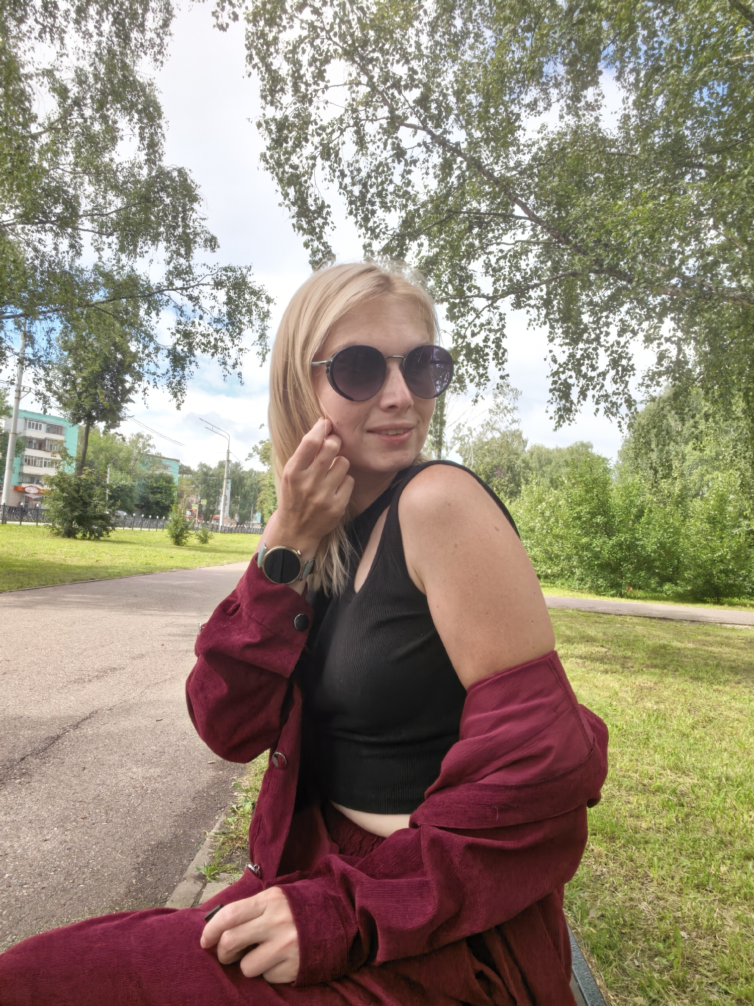 A blonde woman wearing sunglasses, a black crop top, and a maroon jacket and pants sits on a park bench. She is outdoors in a park with trees and grass. In the background, there are apartment buildings and a street. The location is Sterlitamak, Russia.