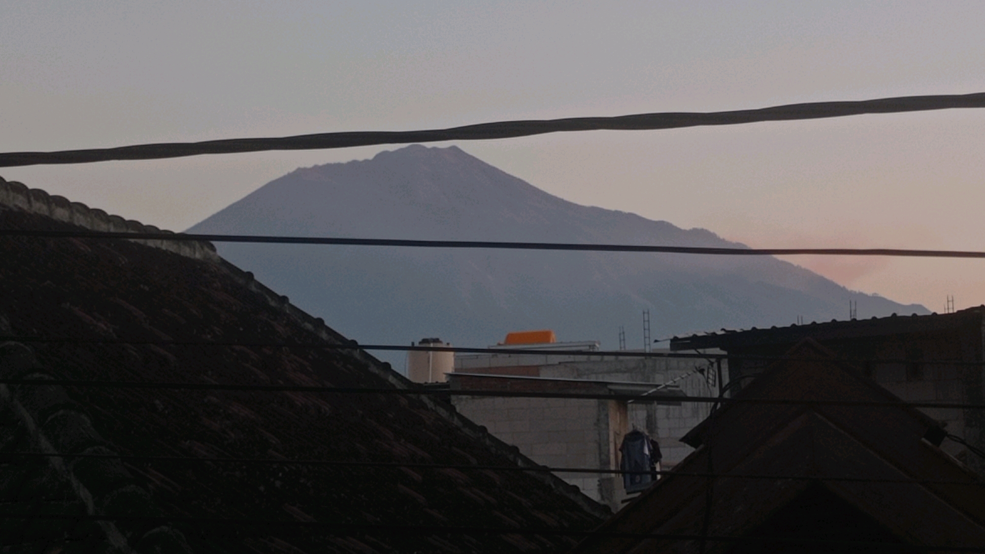 A dimly lit, horizontal medium shot captures a misty, pale blue mountain range in the background, partially obscured by several dark, horizontal power lines. In the foreground, the angled, dark brown tiled roof of a building dominates the lower left quadrant, its texture rough and weathered. To the right of the roof, several other structures are visible, including what appears to be a white water tank, a building with an orange rectangular object on its roof, and other rooftops of varying heights. A few vertical poles, possibly ladders or antenna supports, are also present on these structures. In the mid-ground, a dark blue garment hangs on a clothesline between two buildings. The overall scene suggests an outdoor view from a residential area, possibly at dawn or dusk, with the natural landscape of the mountain juxtaposed against the urban elements.