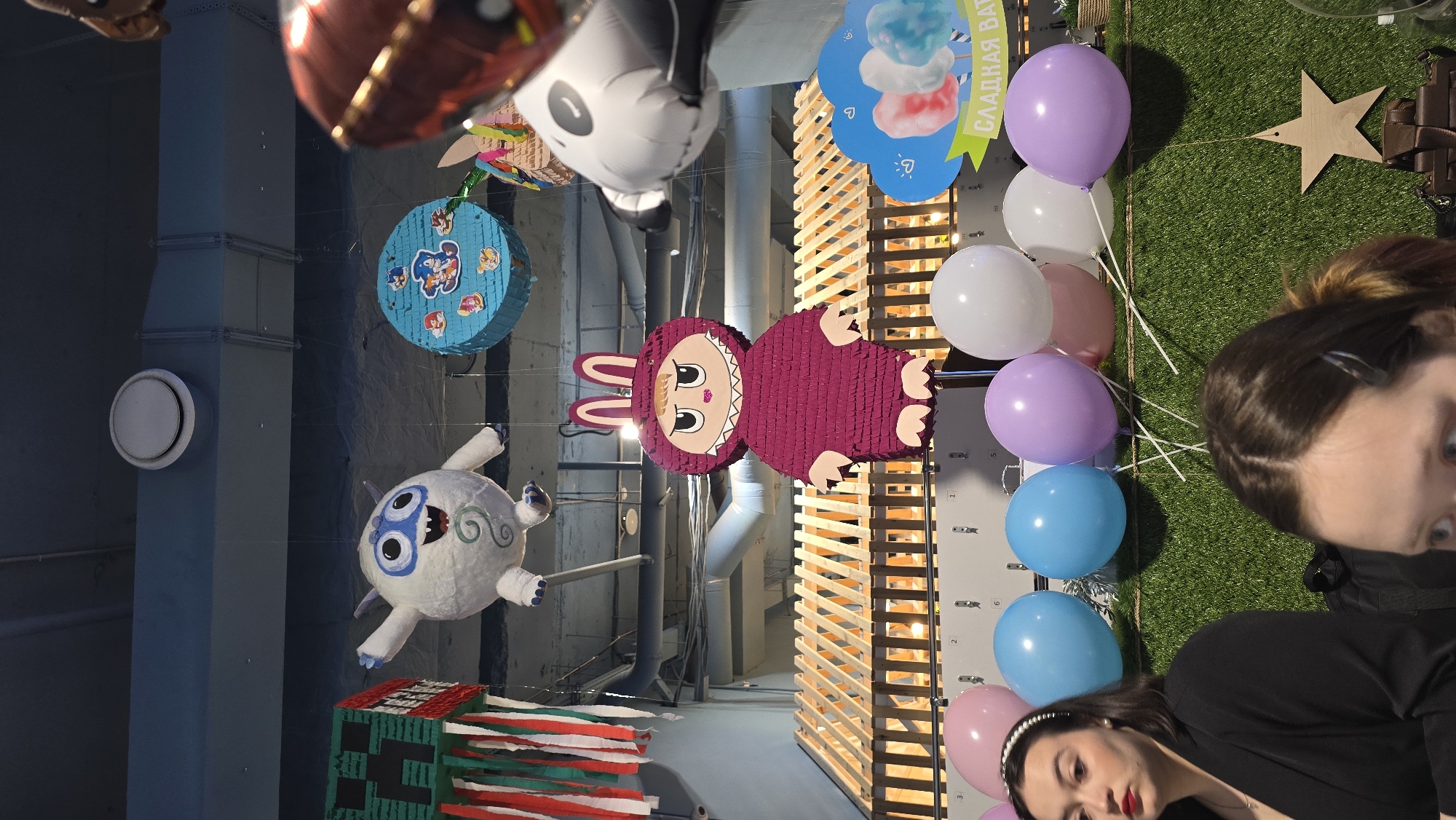 The image depicts an indoor event space, likely for a party or celebration, with various decorations and two partially visible individuals in the foreground. The location is an unknown city in an unknown country.
Suspended from the ceiling, which features exposed blue ventilation ducts and a circular white vent, are several pinatas and balloons. From left to right, a green and red Minecraft Creeper-themed pinata hangs with paper streamers. To its right, a white spherical pinata resembling a yeti or monster with large blue eyes and a wide open mouth is visible. Further right, a round blue pinata adorned with Sonic the Hedgehog characters is present. A large magenta-colored pinata shaped like a character with bunny ears, small eyes, and a prominent mouth is centrally located in the mid-ground. To the far right, a partial view of a black and white inflated object, possibly a panda-like balloon or pinata, is visible.
Below the suspended decorations, a raised wooden slatted structure, resembling a railing or a decorative partition, spans horizontally across the mid-ground.
In the foreground, a vibrant display of balloons in pink, light blue, purple, and white are arranged against a wall covered in artificial green grass. Above these balloons, a blue sign with cloud shapes and images of pink, red, and blue cotton candy is visible, bearing the Cyrillic text "СЛАДКАЯ ВАТА" (SLADKAYA VATA), which translates to "COTTON CANDY". To the right of the balloons, a wooden star decoration is affixed to the grass wall.
Two women are visible in the lower portion of the frame. On the left, a woman with dark hair, wearing a white pearl headband and red lipstick, is partially shown, facing slightly to the right with a neutral expression. In the bottom-center foreground, the top of another woman's head with brown hair is visible.