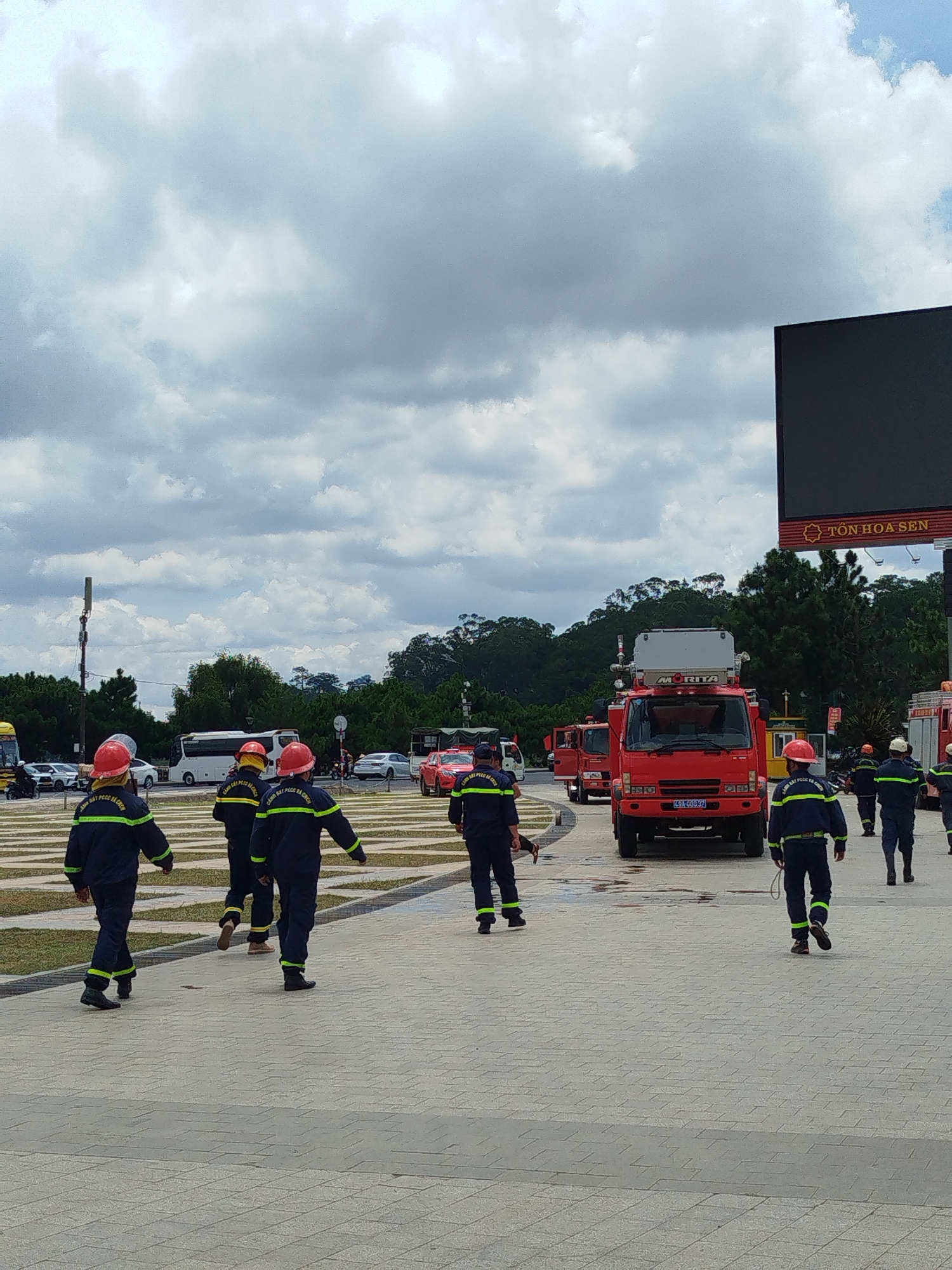 In Da Lat, Vietnam, under a cloudy sky, a group of firefighters in dark blue uniforms with yellow reflective stripes and red helmets are walking across a paved outdoor area. They are positioned in the foreground and midground of the image, walking in various directions. Behind them, several red fire trucks are parked, some with their doors open. One fire truck in the midground has the brand "MORITA" visible on its equipment. In the background, there are trees and a large black screen or billboard with text that appears to read "TON HOA SEN". The scene suggests a potential incident or training exercise involving emergency services in the city.