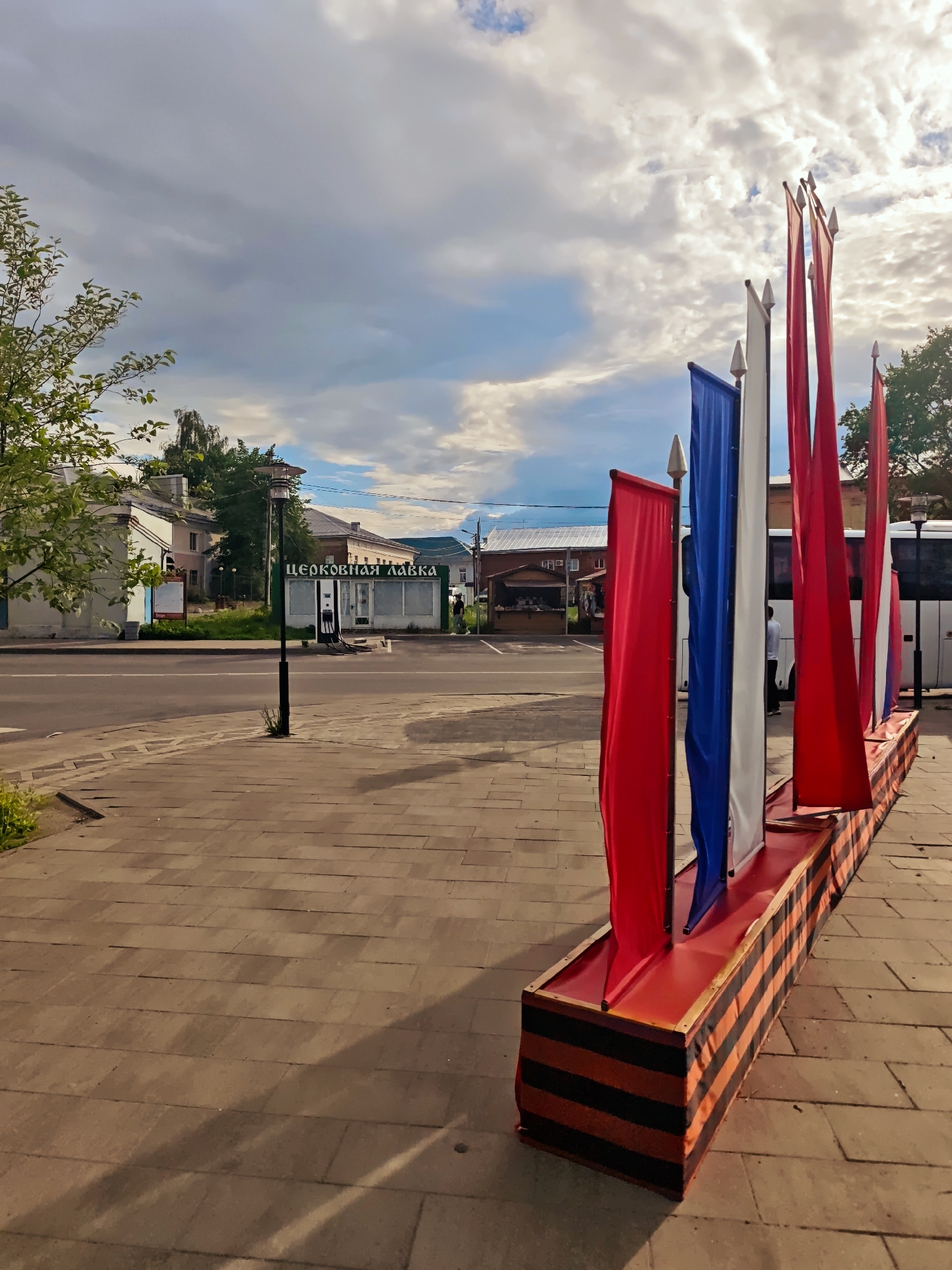 The image shows a street scene in an urban area. On the right side of the frame is a display of vertical flags in the colors red, white, and blue, mounted on a long, low, rectangular base decorated with black and orange stripes. The flags appear to be made of fabric and are slightly ruffled, suggesting a gentle breeze.
In the background, across the street, there is a small building with a sign in Cyrillic script. To its left, there is a lamp post and additional buildings in the distance. A street runs horizontally across the middle of the frame, separating the flags from the buildings across the street. A white bus is parked near the flags.
The foreground is filled with a paved walkway made of rectangular stones. The sky is visible above, with a mix of blue sky and white and gray clouds.