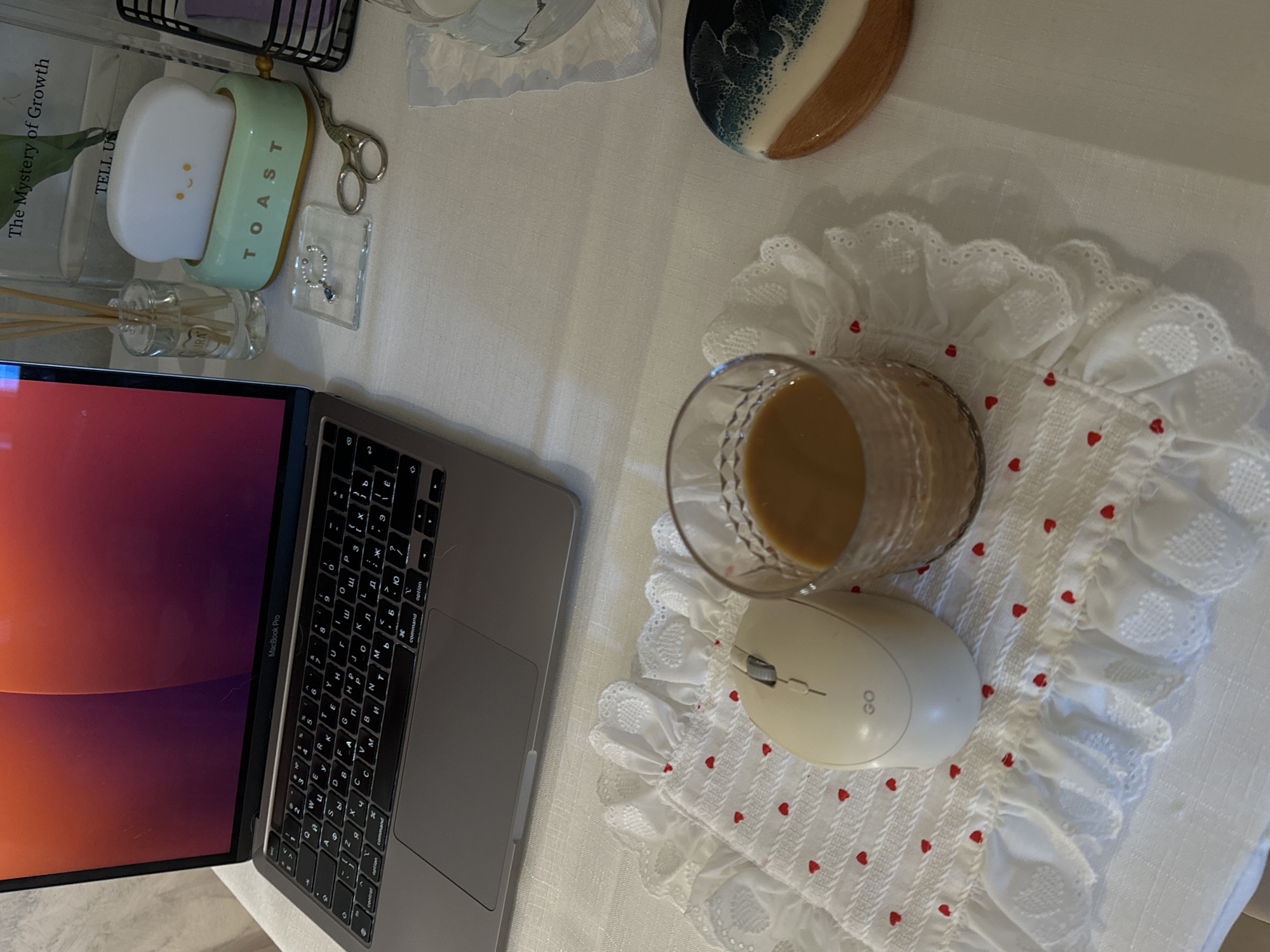 This overhead shot captures a cozy desk or workspace setup, likely indoors. The main subjects are a silver MacBook Pro laptop and various personal and decorative items arranged on a light-colored tablecloth.
An open MacBook Pro occupies the left side of the frame, displaying a vibrant red and orange desktop background. Its keyboard features a Cyrillic layout. To the left and behind the laptop, a clear glass diffuser bottle with wooden reeds is visible. Behind that, a book titled "The Mystery of Growth" with "TELL US" printed below it stands upright. Adjacent to the book is a playful white cloud-shaped light with a smiling face, nestled in a mint green stand labeled "TOAST." Next to the light, a pair of small, ornate gold-colored scissors and a clear acrylic tray holding two rings (one with pearls, one with a blue stone) are neatly placed.
In the upper right corner, a circular decorative item with a dark teal and white resin pattern on a wooden base rests near a ruffled white doily. In the foreground on the right, a clear glass filled with a light brown beverage, possibly coffee or tea, sits on a white, ruffled doily patterned with small red hearts. Beside the glass, a cream-colored wireless computer mouse, labeled "GO," completes the active work/relax zone.
The scene suggests a moment of work or leisure. The overall aesthetic is clean, personalized, and inviting. No people are visible. The context provided indicates this scene is in Bar, Montenegro.