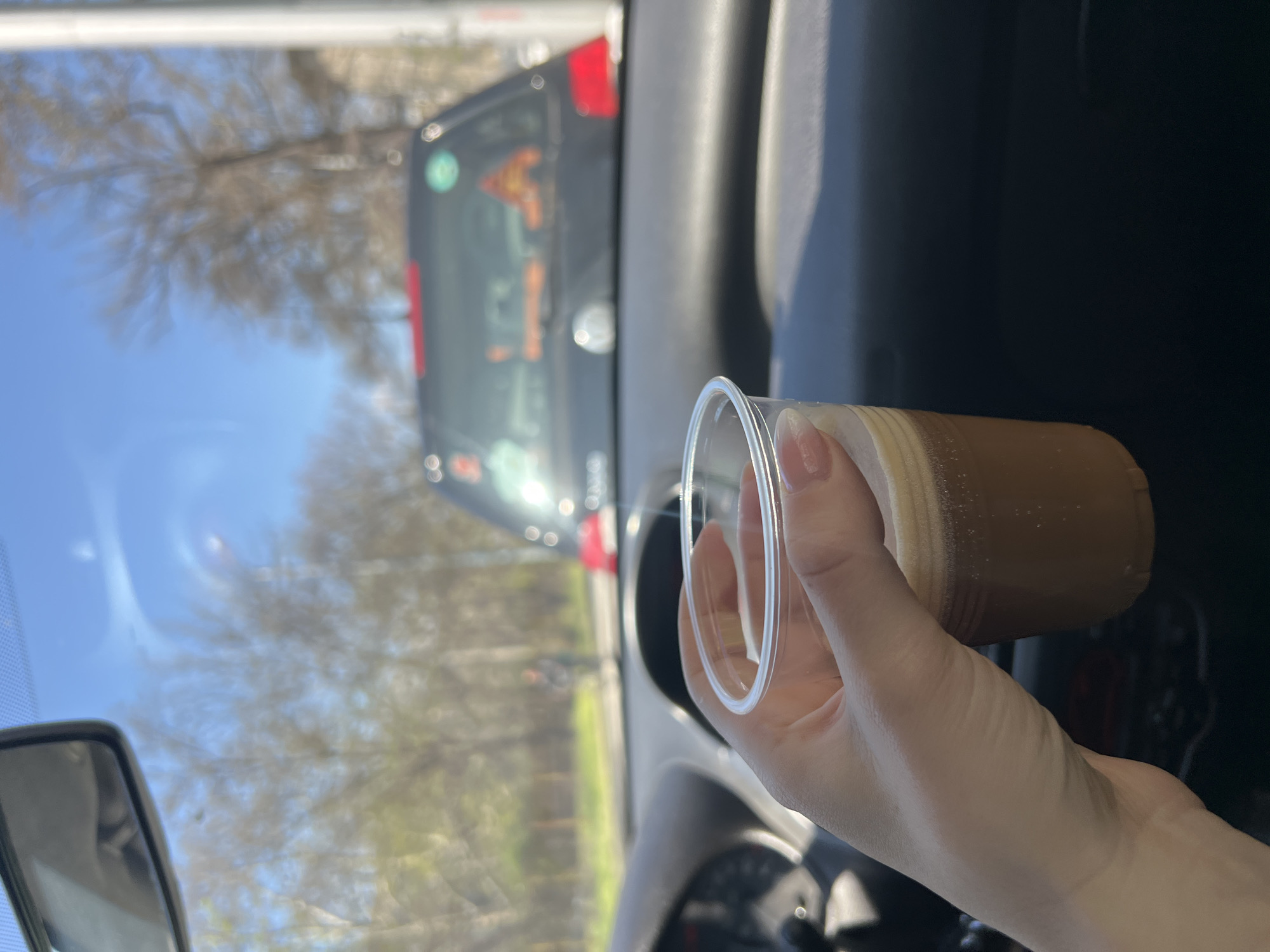 A person is holding a plastic cup with a light brown liquid, likely coffee, while inside a car. The view through the car's windshield shows another vehicle in front, and outside the vehicle, blurred trees are visible under a bright sky. The car appears to be in motion. The location is unidentifiable as Plovdiv, Bulgaria from this image alone.