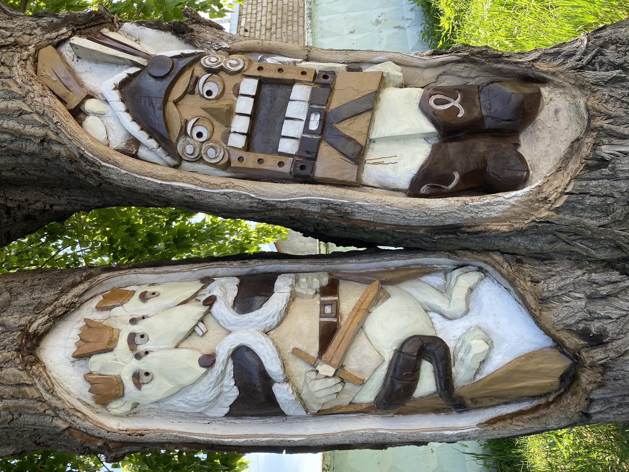 The image shows two wooden carvings on tree branches, likely in an outdoor setting. The top carving depicts a stylized nutcracker figure with a large open mouth and exaggerated features. The figure is rendered in various shades of brown and white.
The bottom carving appears to show a scene with animals, possibly mice or similar creatures, engaged in a battle or confrontation. One figure holds a sword.
The carvings are set into the tree branches, with the bark of the tree forming the natural frame.
Based on the prompt, these carvings are located in the city of Chaikovsky, Russia. The environment around the tree shows foliage and a building in the background.