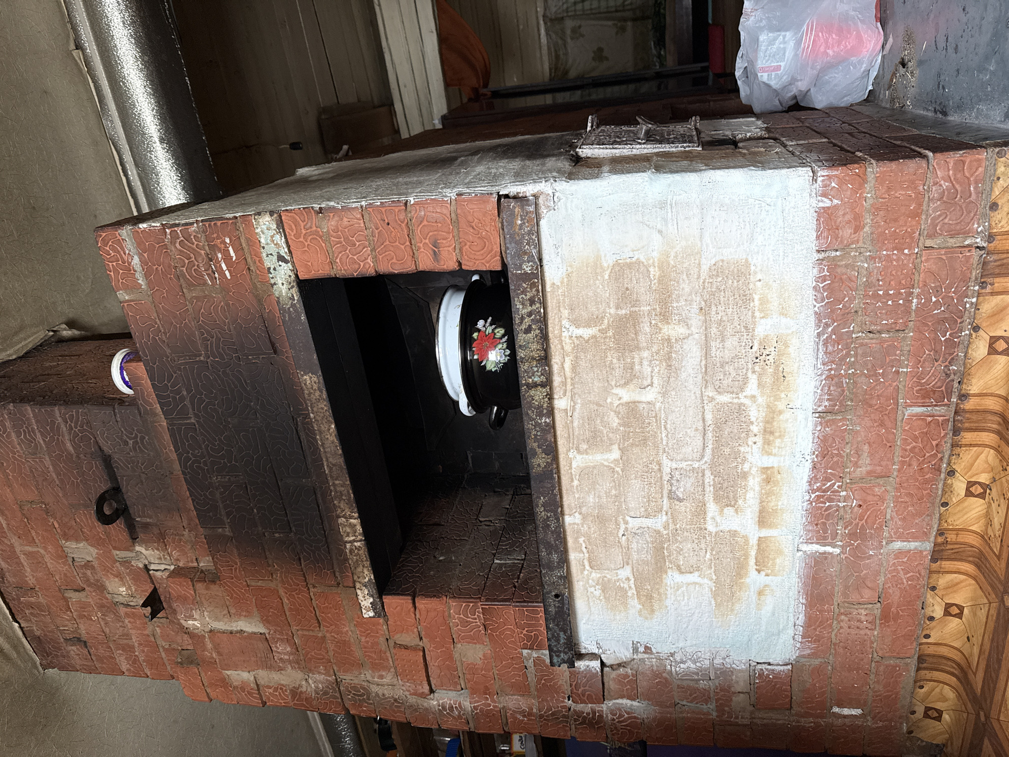 The image shows a brick oven, possibly located in Nizhnyaya Chermoda, Russia. The oven is constructed of red bricks with a textured pattern, with sections either blackened by soot or covered in a faded white coating. The opening of the oven reveals a black interior where a black pot with white trim and a floral design is positioned. The oven top is flat and appears to be made of cement or a similar material. A metal chimney is visible on the left side, suggesting it is a functional oven. The background is somewhat obscured, but suggests an indoor environment with various objects stored nearby.