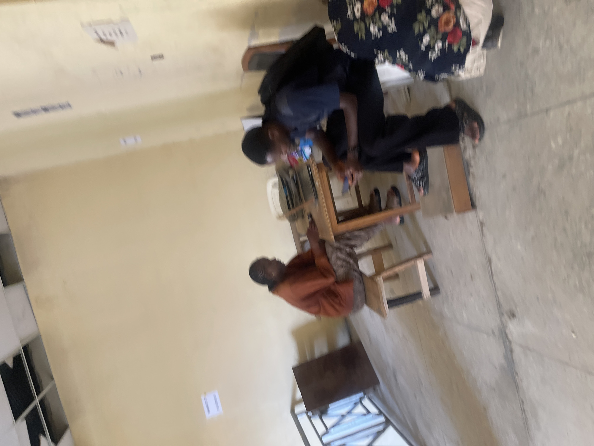 The image depicts an indoor scene, likely in Rafin Par, Nigeria. Two individuals are present, seated. On the left, a person wearing an orange shirt and patterned pants is seated at a wooden table. To the right, another individual wearing a dark shirt, black pants, and a backpack is also seated, looking at a mobile phone. A second table is visible, and a portion of a woman in a floral dress is in the foreground on the right. The room appears to be a waiting area or communal space, with a concrete floor, yellow walls, and a damaged ceiling. A metal railing is visible on the left side of the image. There is a small sign on the wall in the upper left quadrant. The overall lighting suggests an interior environment.