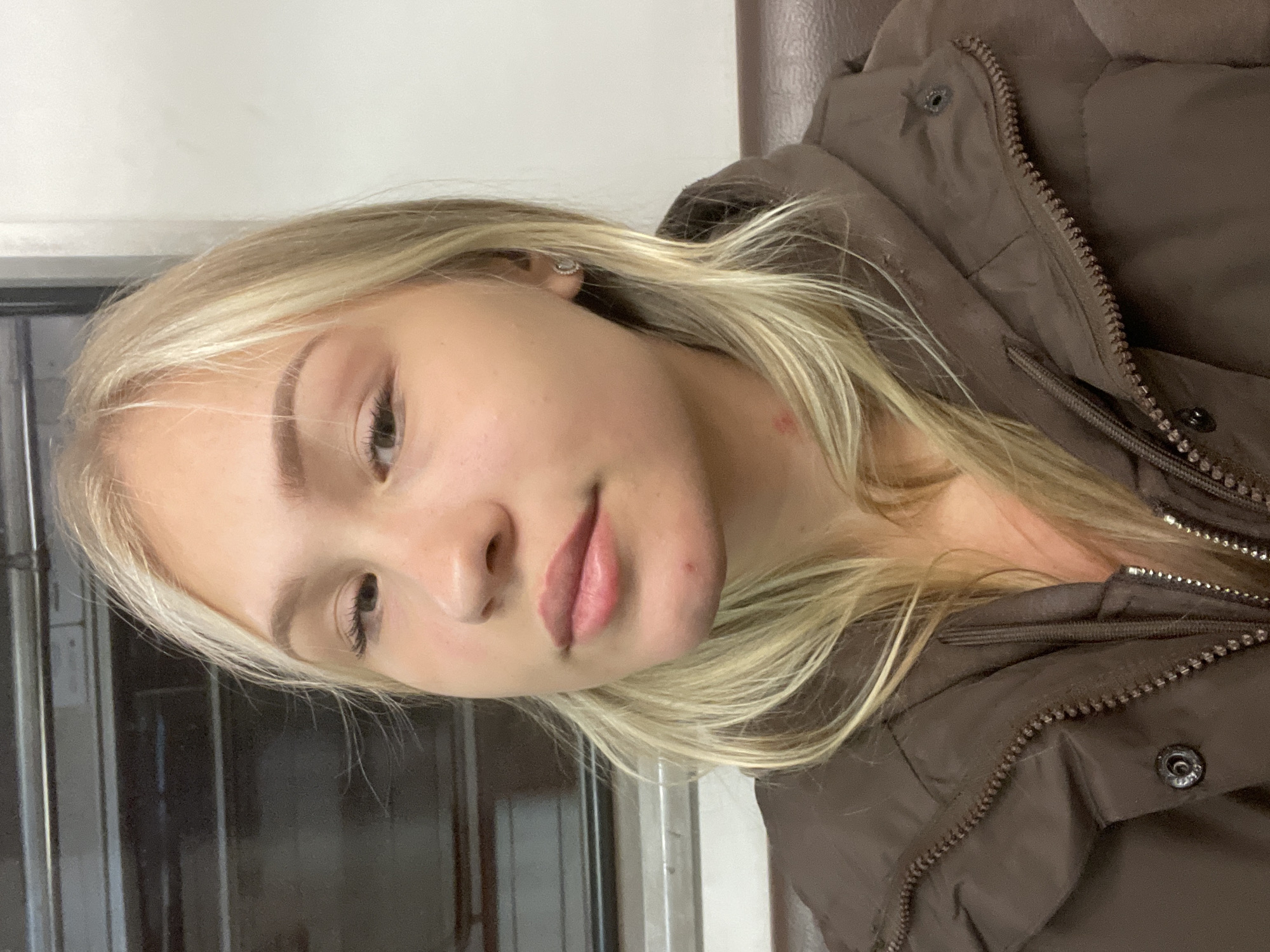 The image shows a young Caucasian woman with blonde hair, light skin, and light-colored eyes. She is wearing a brown puffer jacket. She has a small red mark on her neck and a small blemish on her chin. She is looking at the camera. The background appears to be the interior of a train car. The lighting suggests it is daytime, but it is difficult to ascertain the exact location. It is not possible to determine the location within Moscow, Russia from the provided image.