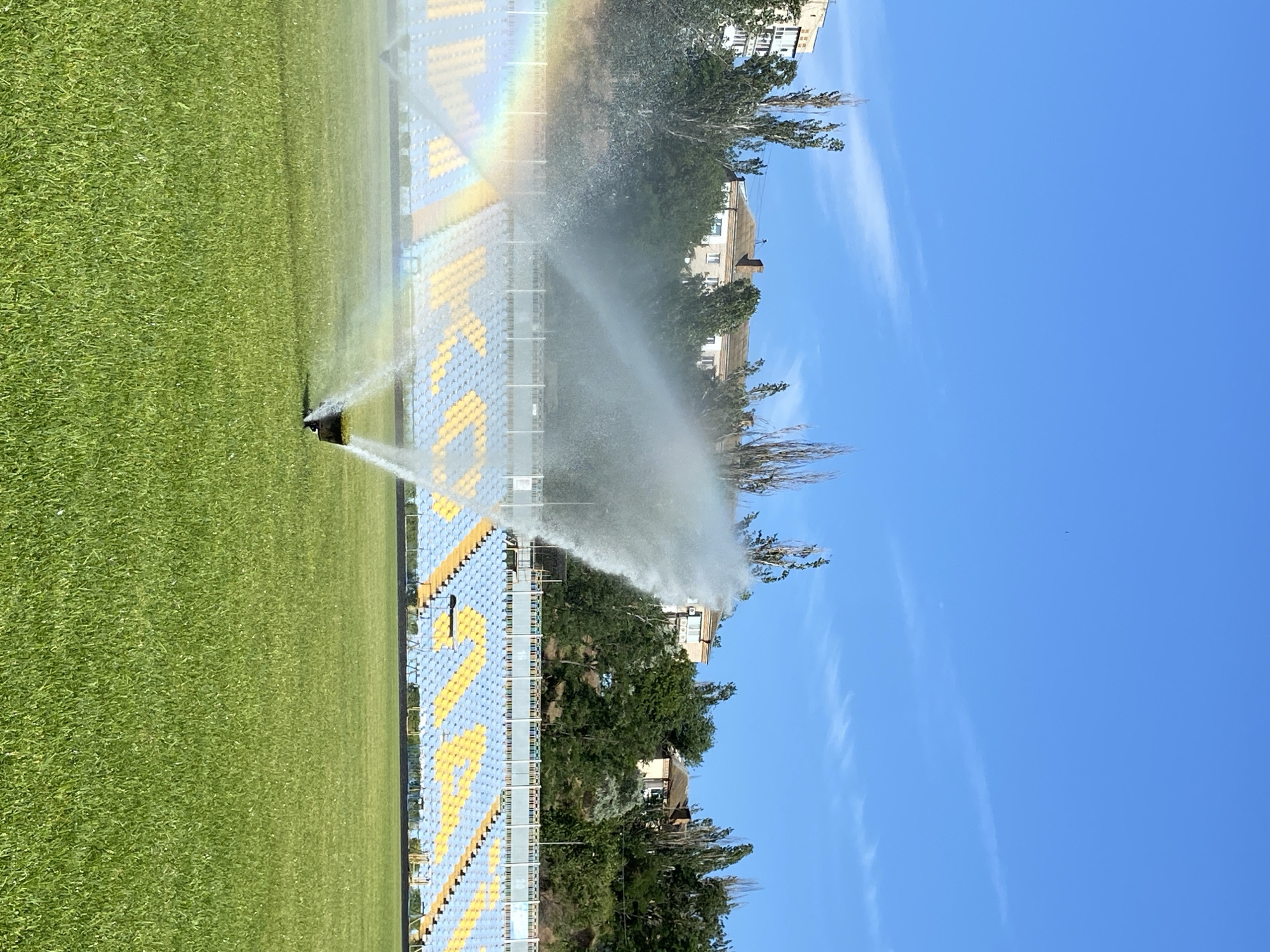 A football stadium's grass field is being watered by two sprinklers, one in the foreground and one further back. A rainbow is visible in the mist of the foreground sprinkler. The stadium seating, in blue and yellow, is visible in the background, with buildings and trees behind it. The sky is clear and blue.