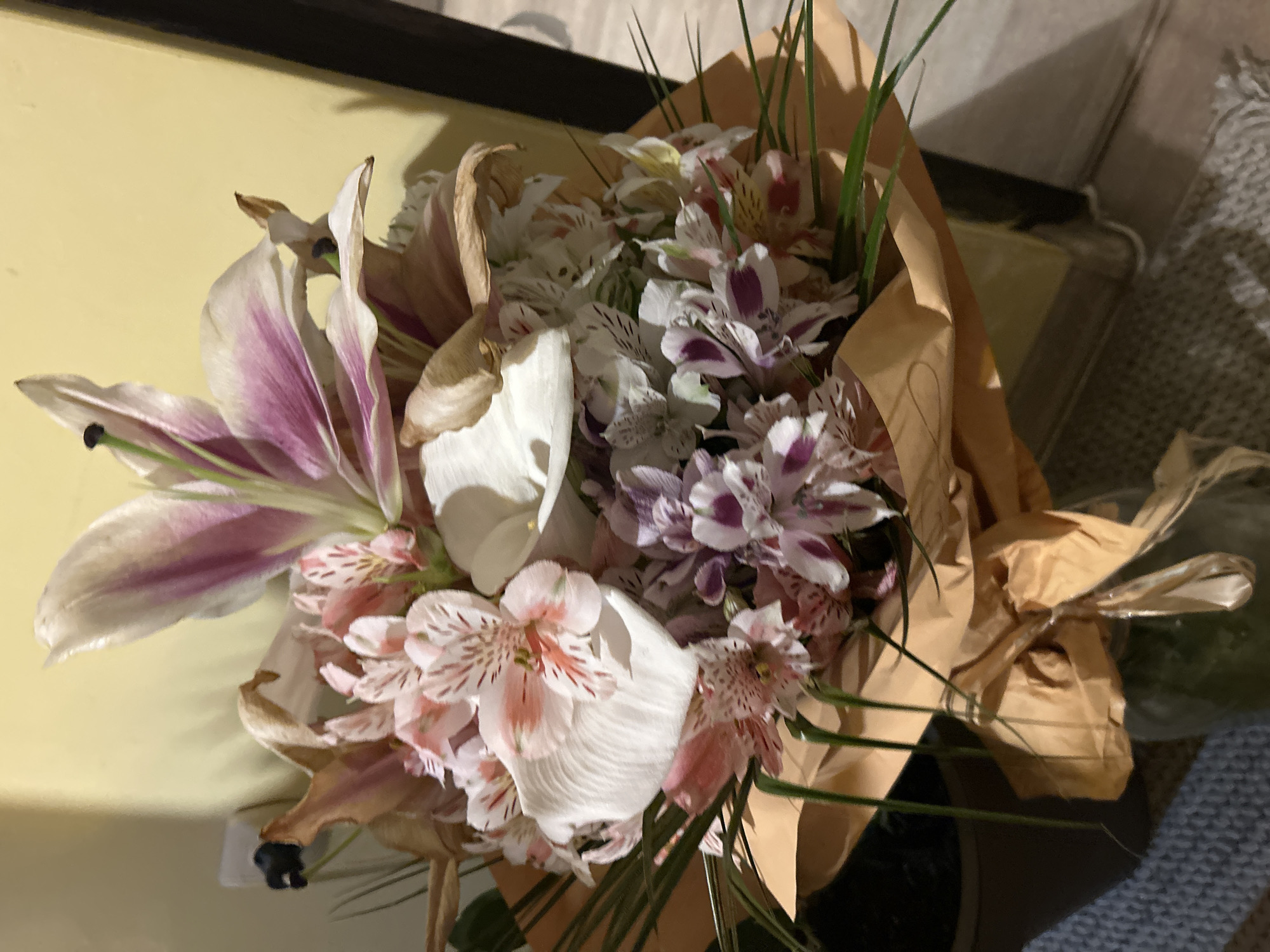 The image shows a bouquet of flowers, including lilies and alstroemeria, wrapped in brown paper. Some of the flowers appear to be wilting or have dried petals. The bouquet is placed indoors, possibly on a surface with a yellow background. The location is not discernable as Burgas, Bulgaria, from the information in the image.