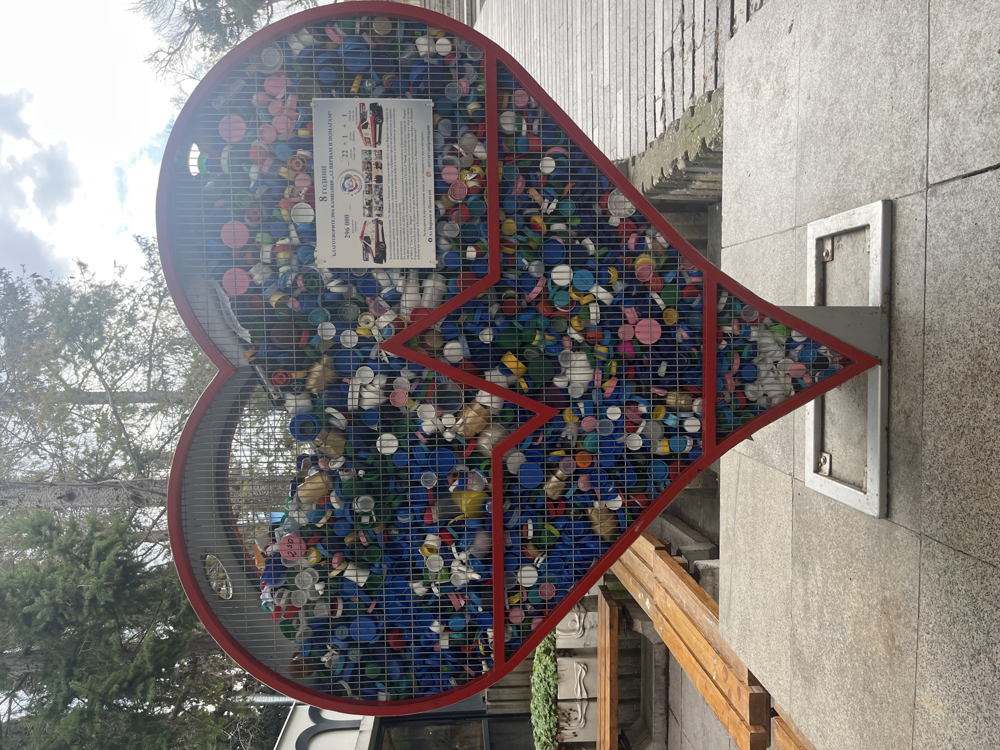 Here's a detailed description of the image:
**Overview:**
The image depicts a large, heart-shaped metal structure filled with colorful plastic bottle caps. The structure appears to be a collection point for recycling, with the collected caps forming the shape of a heart. A sign is attached to the heart structure, displaying information about the charity campaign "I Believe and Help".
**Specific Elements:**
* **Heart Structure:** The structure has a red outline and is filled with a wire grid that contains a dense collection of plastic caps in various colors (primarily blue, white, pink, and yellow).
* **Signage:** The sign contains details about the charity campaign, including the name ("I Believe and Help" - "Az Vyarvam i Pomagam").
* **Surroundings:** The heart structure is positioned outdoors on what appears to be a stone or concrete platform. In the background, there are trees and buildings.
* **Location (based on OCR):** The OCR mentions "MBSAL Sveta Anna Varna", which indicates that the image was taken in Varna, Bulgaria, specifically at or near the Sveta Anna University Hospital.
