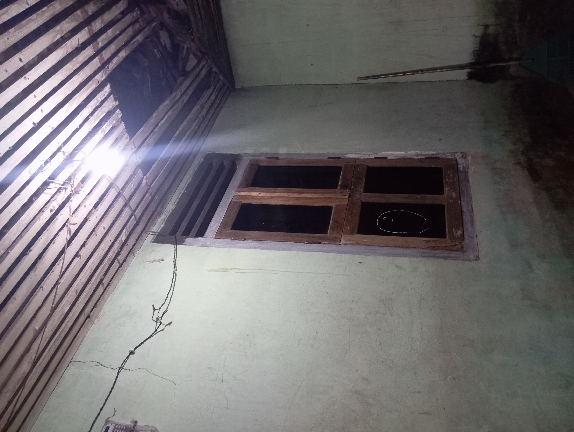 The image depicts the interior of a room, likely a dwelling, in Rejosari, Indonesia. The walls are painted a pale green and show signs of wear, including cracks and some discoloration near the base. Above, the ceiling is constructed of wooden planks, with some sections appearing damaged and revealing darkness beyond. A single light bulb hangs from exposed wiring, illuminating the space.
A prominent feature is a wooden-framed window with multiple panes, some of which are dark or obscured. On the wall to the left of the window, there is a drawing of a face with question marks. To the right of the window, a broom with a green plastic head and a wooden handle stands upright against the wall. The floor is not clearly visible, but the lower portion of the walls suggests a dark, potentially dirty surface. The overall impression is one of a simple, possibly neglected, living space.