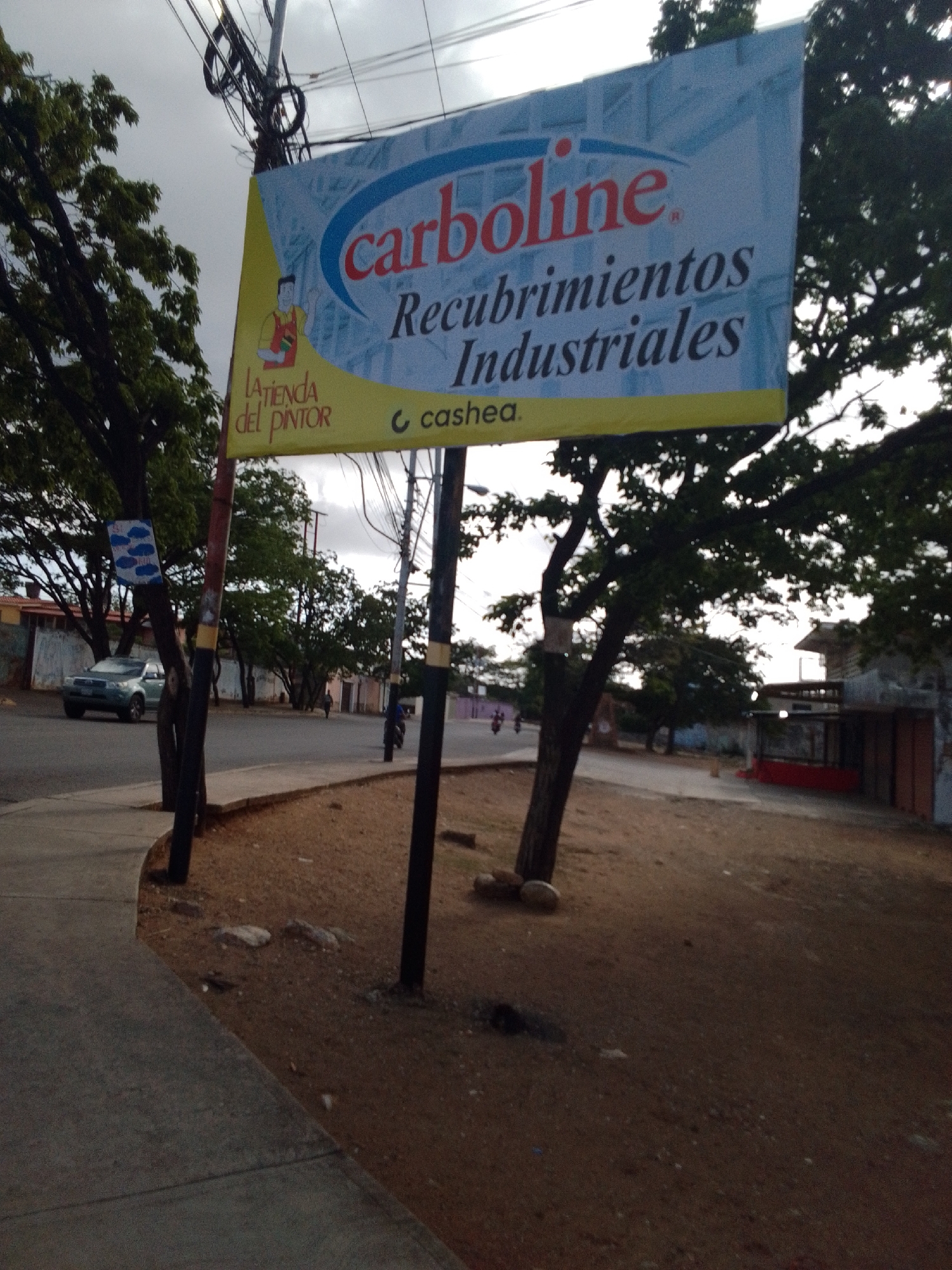 The image depicts an outdoor scene with a prominent billboard advertising "carboline" and "Recubrimientos Industriales". To the left of the billboard, a sign reads "LA TIENDA del PINTOR". A grey sedan is parked on a street to the left, and several trees with lush green foliage are visible throughout the scene. A sidewalk runs along the left side of the image, leading towards a building with a red awning in the background on the right. The environment appears to be a street in Altagracia, Venezuela. The sky is overcast, suggesting a cloudy day.