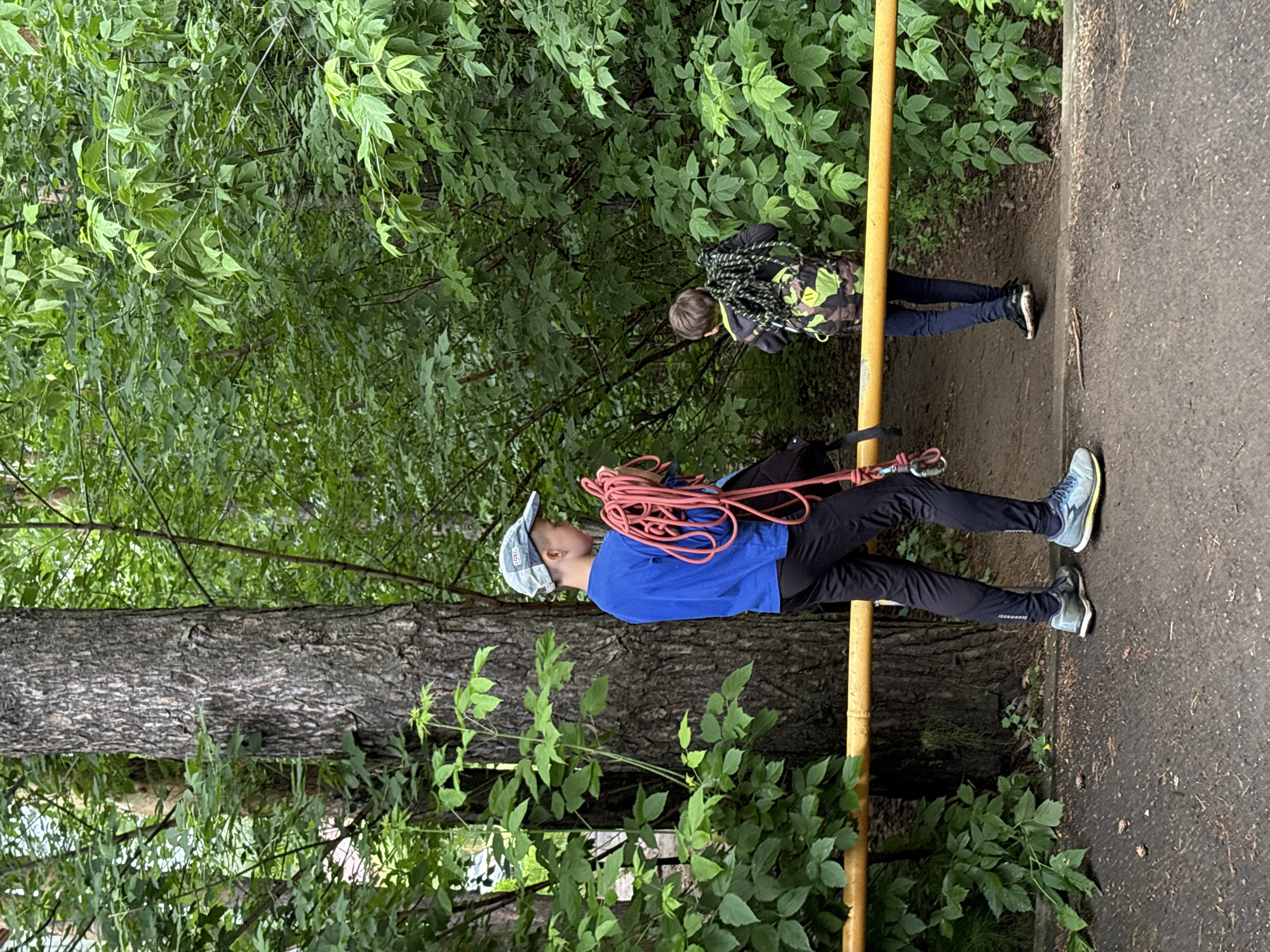 The image depicts two individuals, likely children or teenagers, in a wooded area, possibly part of a park or recreational space in Chaikovsky, Russia. The foreground shows a dirt path with some scattered pinecones, bordered by a yellow horizontal bar.
The individual on the left, facing away from the camera and towards the right, is wearing a blue long-sleeved shirt, black pants, and a light-colored baseball cap. They are holding a thick, bright red rope coiled in their arms. Their stance suggests they are either preparing to move or have just stopped.
The second individual, further to the right and also facing away, is wearing a camouflage-patterned backpack with green and brown hues. They appear to be carrying a coiled rope, darker in color than the one held by the first person, on their back. They are wearing dark pants and dark shoes.
The background is dominated by dense green foliage, indicating a natural environment with numerous trees and bushes. The light suggests it is daytime, with overcast conditions or shade from the trees. The overall scene suggests an outdoor activity, potentially related to climbing or an adventure program, given the presence of ropes and the natural setting. There is no specific indication of urban infrastructure or recognizable landmarks that would pinpoint the exact location within Chaikovsky, beyond it being a green, wooded area.