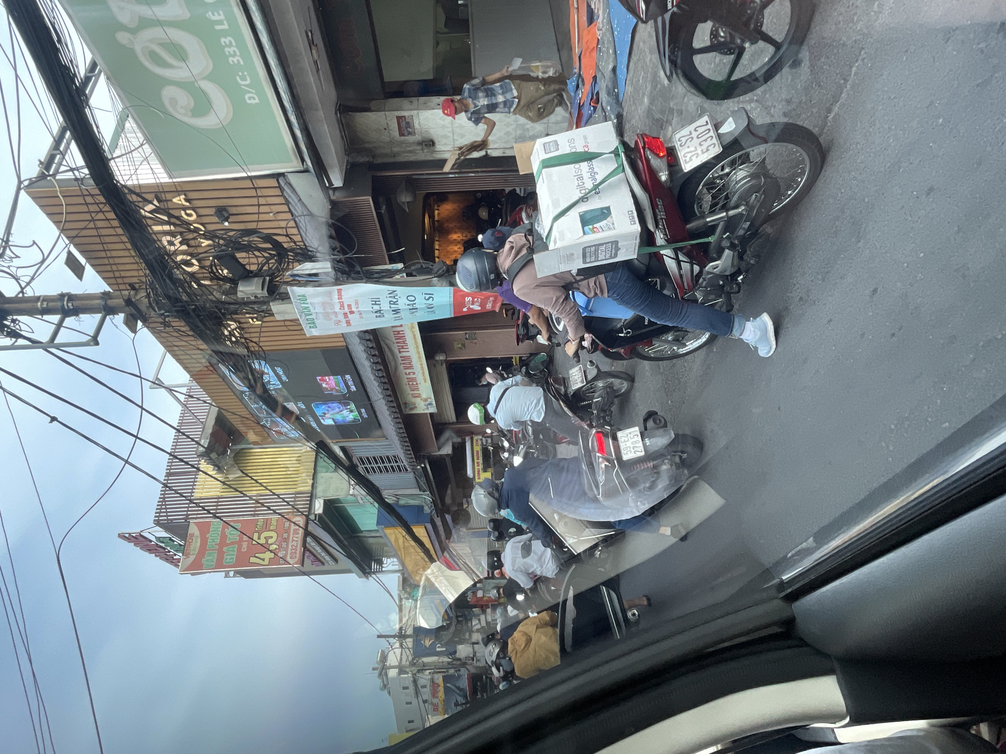 This image captures a busy street scene in Thủ Đức City, Vietnam, likely during daylight hours, with a moderate amount of traffic congestion. The perspective is from inside a vehicle, looking out through the windshield. The road is asphalt, and there are several motorcycles and scooters, many with riders wearing helmets. Some motorcycles are carrying large boxes or packages.
Along the street, there are buildings with various signs and awnings. One prominent sign, partially obscured, appears to advertise "VIETTEL," a telecommunications company. Another sign reads "VICTORIA YOGA." Green signage with Vietnamese text and a red letter "C" is visible on the right side of the image, indicating a business. The sky is overcast. The environment suggests a typical urban setting with overhead electrical wires and poles. The general atmosphere is one of everyday urban life and commerce.