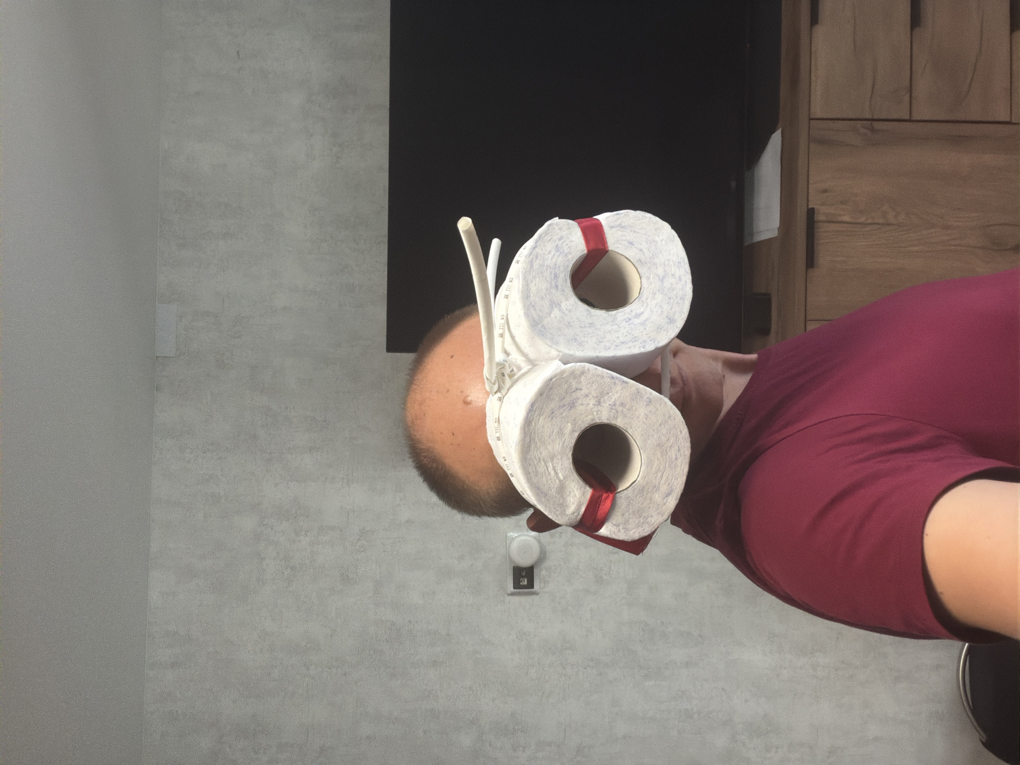A man in a maroon t-shirt stands indoors, facing left, with two rolls of toilet paper strapped to his head as makeshift goggles. A red ribbon is tied around each roll. He has a bald head. The background includes a grey textured wall with a light switch and a dark television screen above a wooden cabinet with drawers. The image is likely taken in a residential setting in Yekaterinburg, Russia.