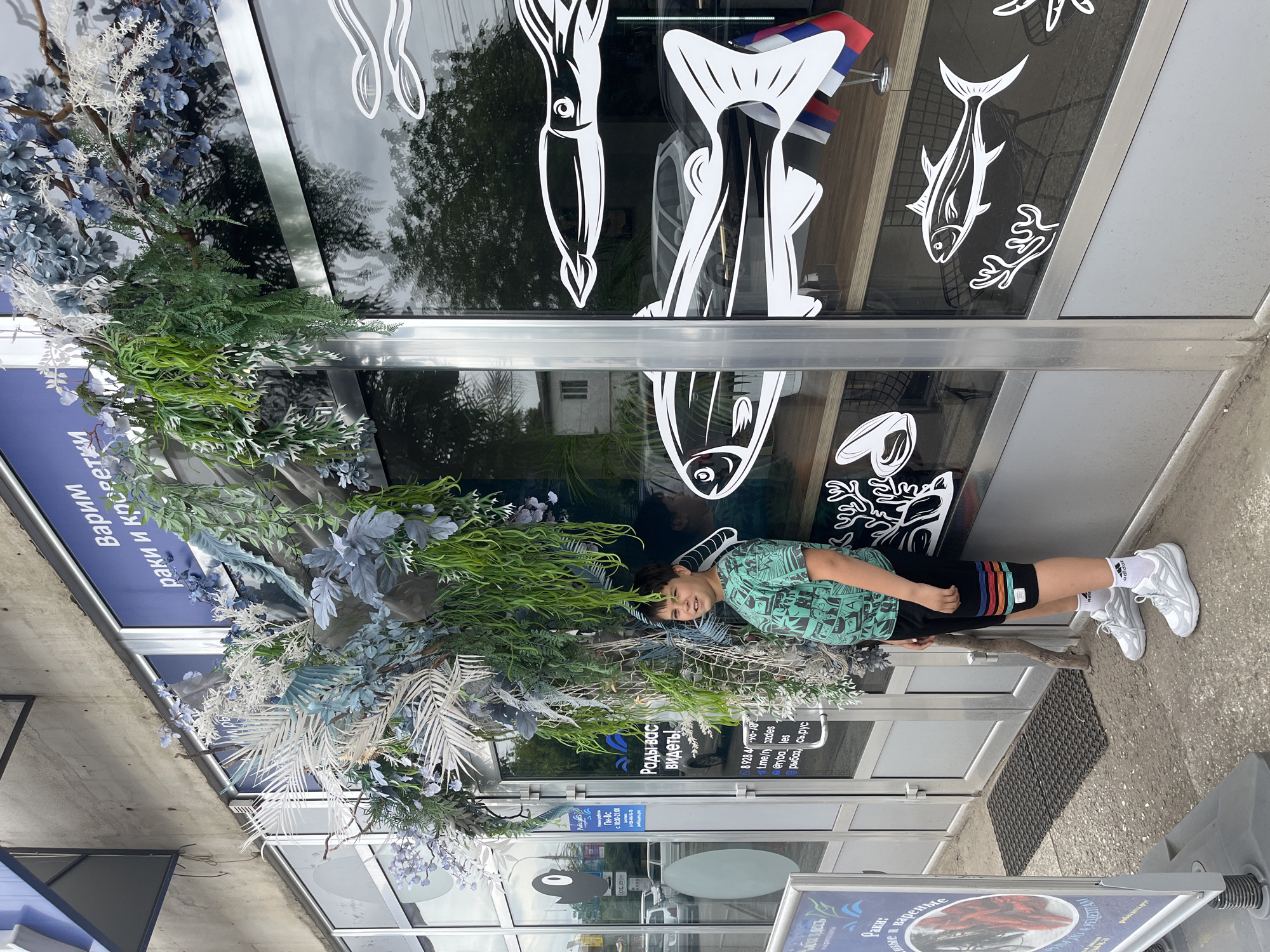 A young boy stands outside a storefront in Krasnodar, Russia, posing for a photo. The store appears to be a seafood market, as evidenced by the decals of fish and sea creatures on the large glass windows and the sign that reads "Рады вас видеть!" (Glad to see you!). The boy is wearing a green t-shirt with a graphic print, black shorts with colorful stripes, white socks, and white sneakers. He is smiling at the camera. The storefront is decorated with artificial plants and branches, creating an organic, underwater theme. The exterior wall of the building is made of concrete, and the storefront has a metallic frame. Cars and trees are visible through the windows, reflecting the outside environment. A small table with two Russian flags is placed near the right side of the entrance. The ground in front of the store is paved with asphalt and concrete.