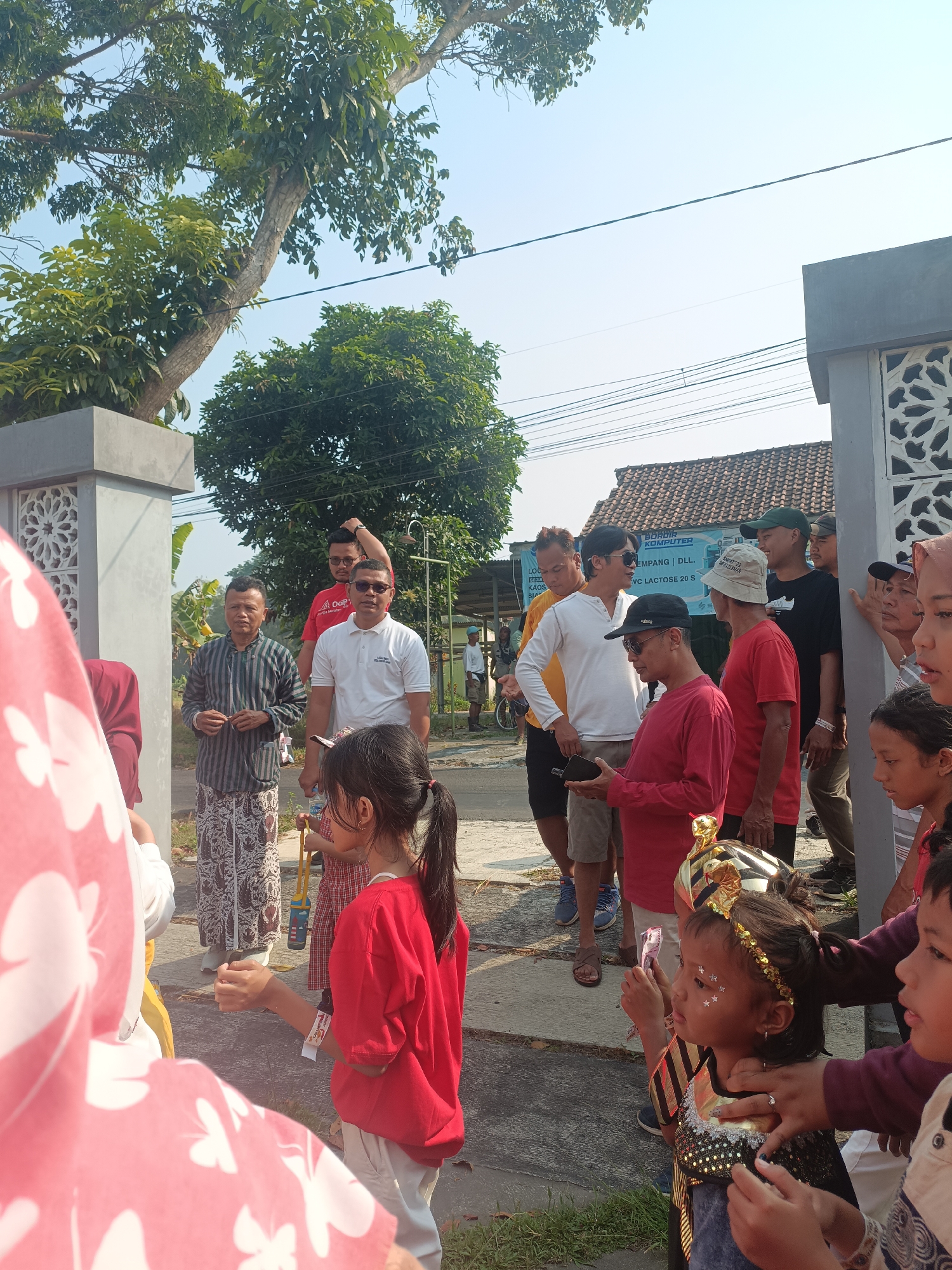 In the city of Sleman, Indonesia, a group of people, including children and adults, are gathered outdoors near a building and trees. The scene appears to be a community event or celebration.
Several individuals are dressed in traditional Indonesian clothing, such as sarongs. Some are wearing casual attire, including t-shirts and hats. A few children are dressed in costumes, one with a gold headdress and sequins, and another in a red shirt.
The environment suggests a sunny day with a clear sky. There are buildings with tiled roofs in the background, along with power lines. The ground is paved with asphalt and some grass is visible.
The people are interacting with each other, with some appearing to be observing or participating in an activity. One adult is holding what looks like a trophy. Another person is holding a small blue container. Some are looking towards the front, while others are conversing. The overall atmosphere seems positive and engaging.