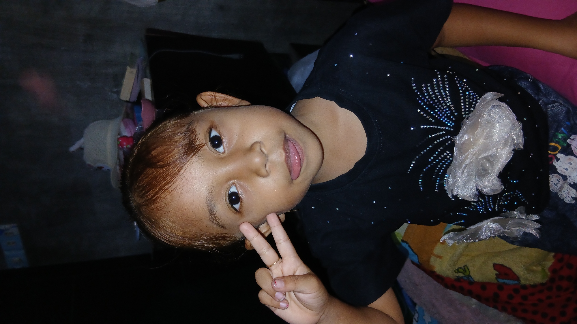 This media file shows a close-up, vertically oriented shot of a young child, likely a girl, making a peace sign with her right hand. She has brown hair, brown eyes, and fair skin. She is wearing a black t-shirt adorned with sparkly embellishments that form a sunburst or fireworks pattern. A piece of translucent plastic is attached to the shirt in front of the pattern. The child's tongue is slightly sticking out.
The background is dark and somewhat cluttered, suggesting an indoor setting. To the left of the child, some items are visible, including what appears to be a straw hat, books, and other colorful objects, possibly toys or accessories. The setting appears to be a domestic environment, possibly a bedroom, given the presence of bedding and a pink surface in the upper right corner. The lighting is dim, indicating it might be nighttime or an indoor space with low light. No specific time of day or weather conditions can be determined.
The child's expression is playful and engaging, with her eyes looking directly at the camera and her peace sign gesture conveying a sense of lightheartedness. There is no visible text in the media file. The location context of Keudejungkagajah, Indonesia, provides a geographical reference for the scene.