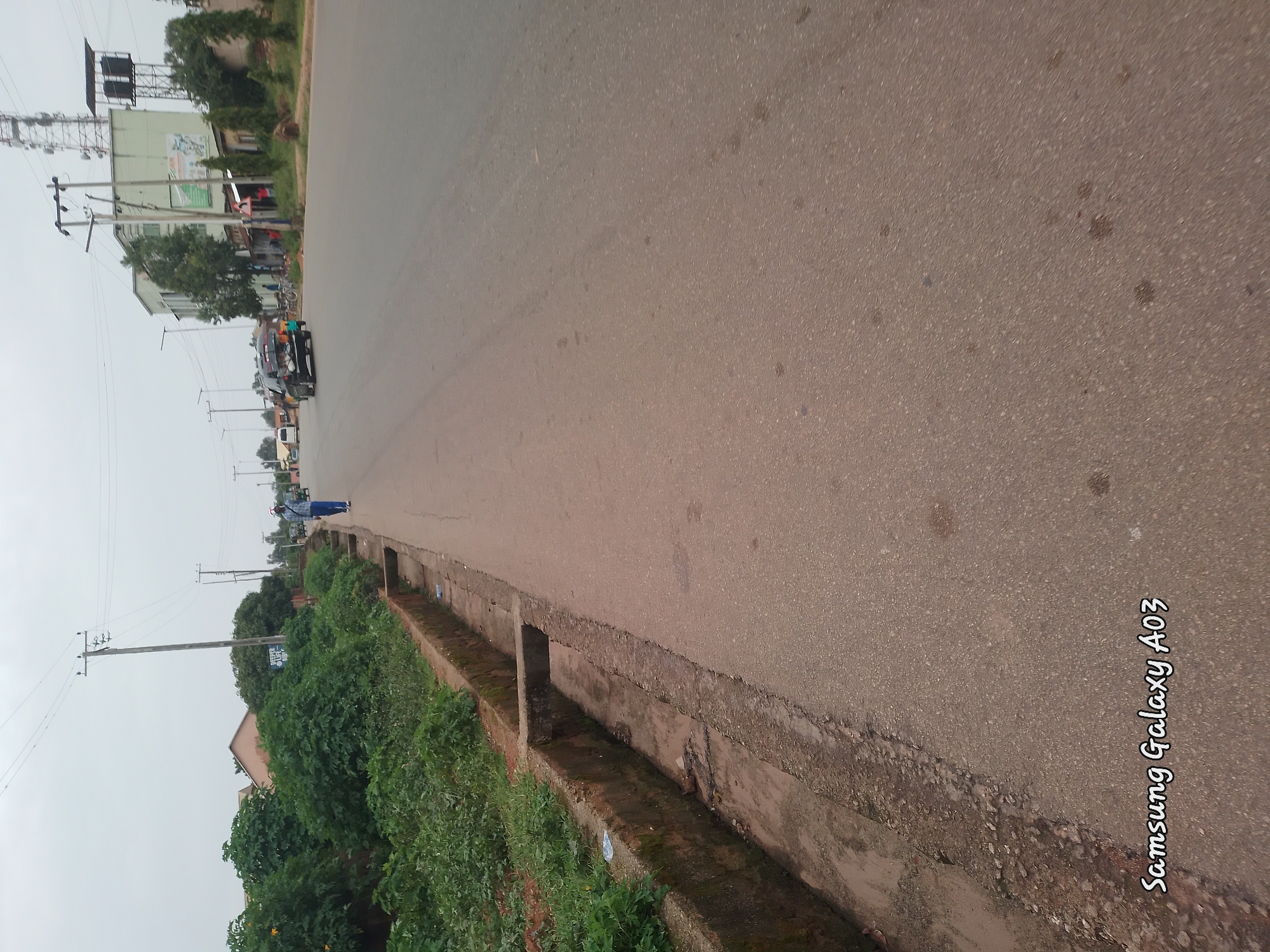 The image depicts a street scene in Jos, Nigeria, on an overcast day. The road is asphalt, with a concrete drainage ditch running along the left side of the image. Dense green vegetation borders the ditch. A person in blue pants and a plaid shirt walks on the right side of the road. In the distance, several vehicles, including a black SUV and a white van, are visible. Buildings and a tall telecommunications tower are also present in the background. The overall impression is of a moderately busy urban street.