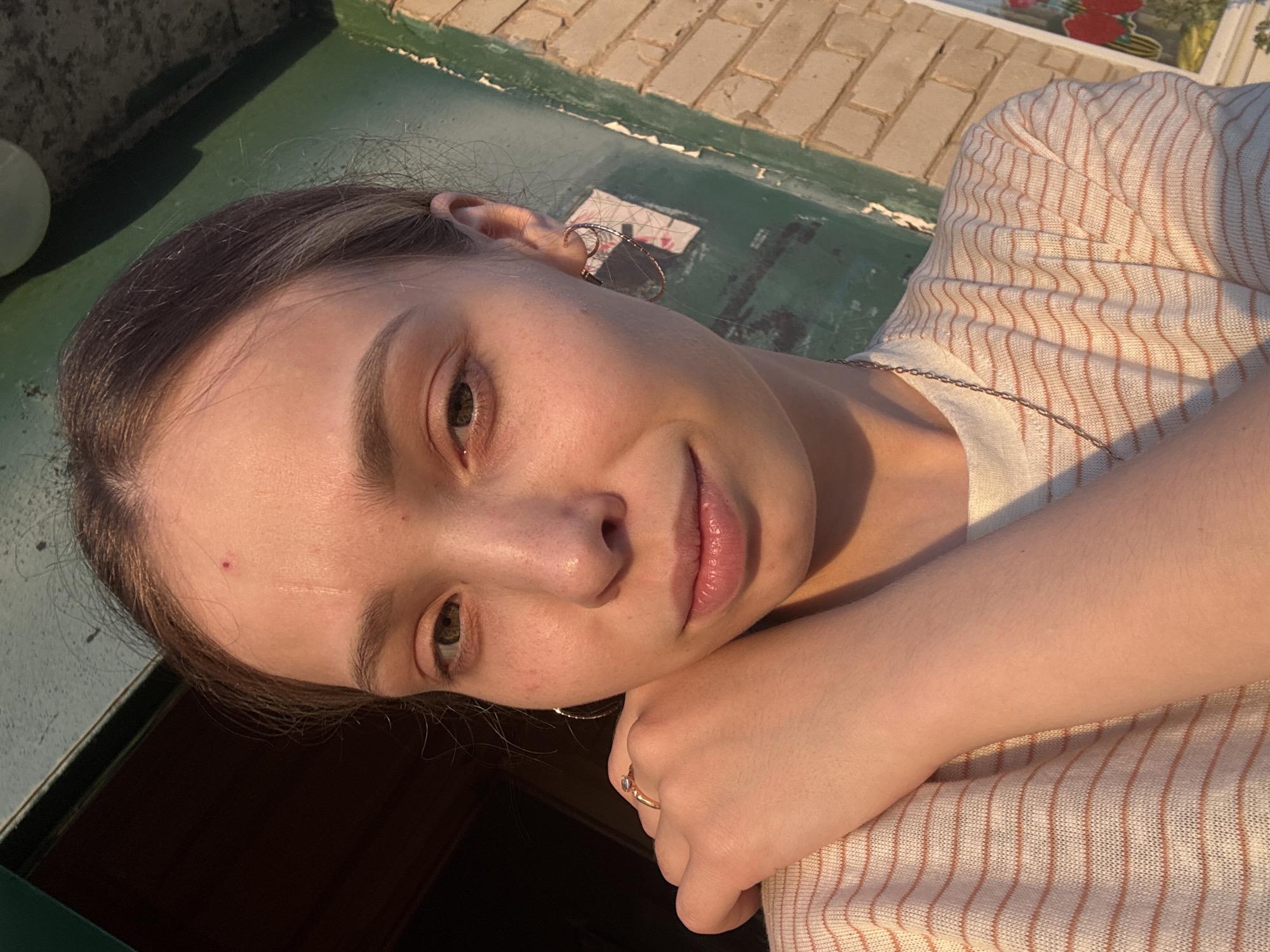 A young woman is facing the camera with a slight smile, resting her head on her hand. She has fair skin, brown hair, and brown eyes. She is wearing earrings and a ring, as well as a light-colored, striped shirt. The background shows brickwork and green painted surfaces, suggesting an outdoor setting, possibly in Кемуль, Russia, based on the image's origin. The lighting indicates it is daytime.