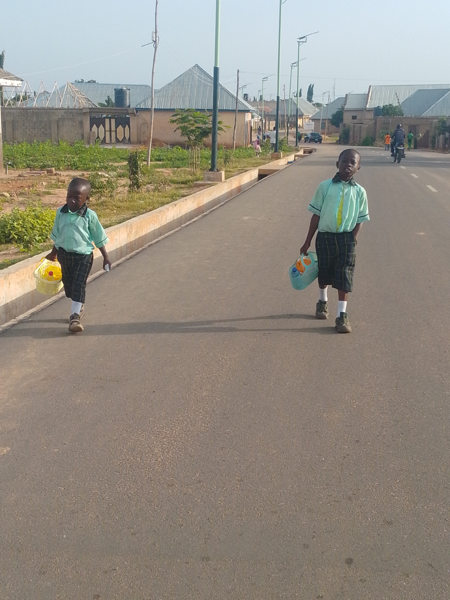 In Tempure, Nigeria, two young boys in school uniforms walk down a paved road. The boy on the left wears a light blue shirt and checkered shorts, carrying a yellow bucket in his right hand. The boy on the right wears a similar light blue shirt and checkered shorts, carrying a light blue basket in his left hand. Both boys are wearing white socks and dark shoes. The road is bordered by a concrete curb and a strip of vegetation. In the background, there are houses with corrugated metal roofs, some with fences and trees. Other people and vehicles are visible in the distance, indicating a residential or community area. The sky is clear and the lighting suggests it is daytime.