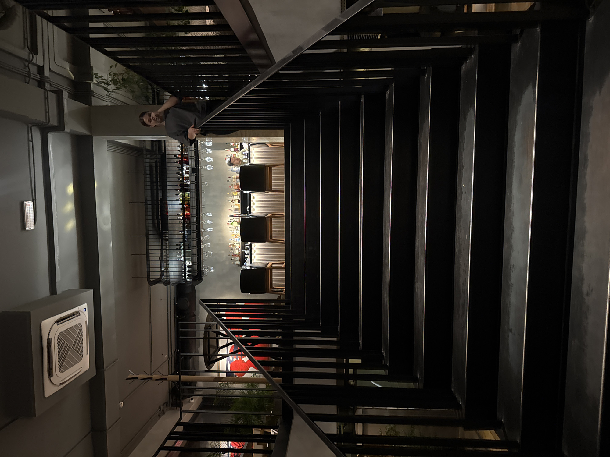 An indoor shot shows a dark, modern staircase leading up to a bar area. The staircase is made of black metal with wooden steps. At the top of the stairs, a bar is visible with bottles lining the shelves. A bartender, wearing a gray shirt, is standing behind the bar. Another person, also wearing a gray shirt, is leaning on the railing near the top of the stairs. The environment appears to be a restaurant or bar with dim lighting.