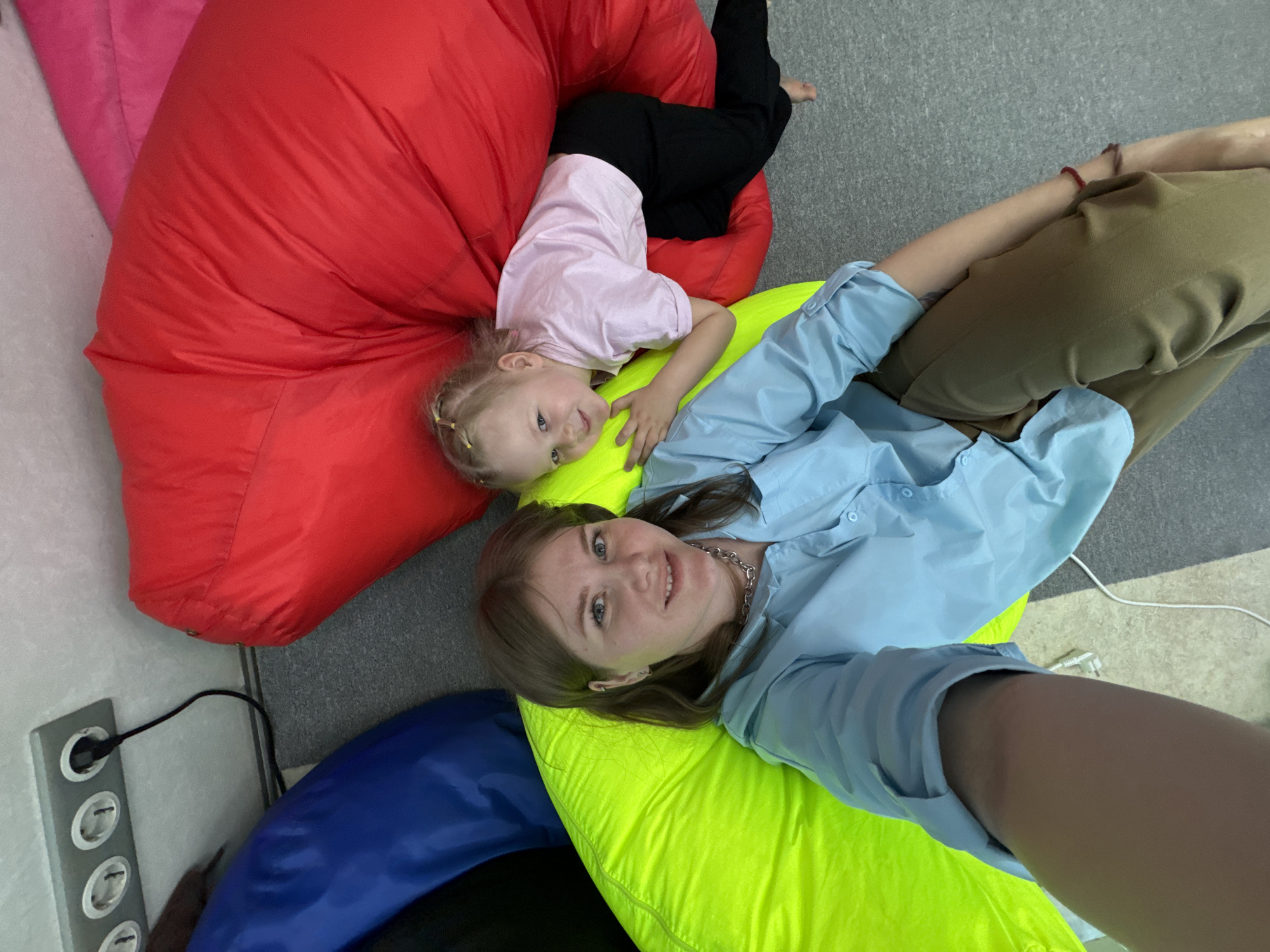 This indoor image, taken in Perm, Russia, depicts two females, an adult and a young child, relaxing on colorful bean bag chairs. The scene is captured from a high-angle, looking down at them as they recline and smile at the camera.
In the foreground, an adult woman with light brown hair and blue eyes lies on a bright neon yellow bean bag chair. She wears a light blue button-up shirt, a silver chain necklace, and olive-green pants. She is smiling directly at the viewer, suggesting a selfie.
Behind her, on a large red bean bag chair, a young child with blonde hair is playfully positioned upside down, her head near the adult's. She wears a light pink t-shirt and black pants and also smiles at the camera. Her feet are visible at the top left of the red bean bag.
The floor is covered with a grey carpet. In the bottom left corner, a silver power strip is mounted on a light-colored wall, with a black power cord plugged into one of its sockets. Part of a blue bean bag chair is visible in the lower left. Another white cord is seen on the carpet near the adult's right arm.
The overall atmosphere is cheerful and relaxed, indicating a moment of comfort and connection. The setting appears to be a casual lounge or play area during the daytime. There is no visible text in the image.