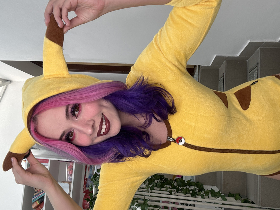 A young adult female is depicted indoors, centered and smiling directly at the camera. She is wearing a yellow, plush long-sleeved bodysuit designed to resemble a Pikachu, featuring brown accents on the sides, a brown front zipper, and a Poké Ball emblem as the zipper pull. The suit includes a hood with two upright, brown-tipped ears, which she holds with her raised hands. Her long, wavy hair is dyed pink on the upper sections and purple on the lower sections. Her face has full makeup, including red eyeshadow, winged eyeliner, and dark red lipstick. The background on the left side features a partial white shelving unit with books and a white railing with a green climbing vine. On the right, a light-colored wall and a grey staircase with dark rectangular mats on each step are visible. The scene is brightly lit. The location is the City of Zagreb, Croatia.