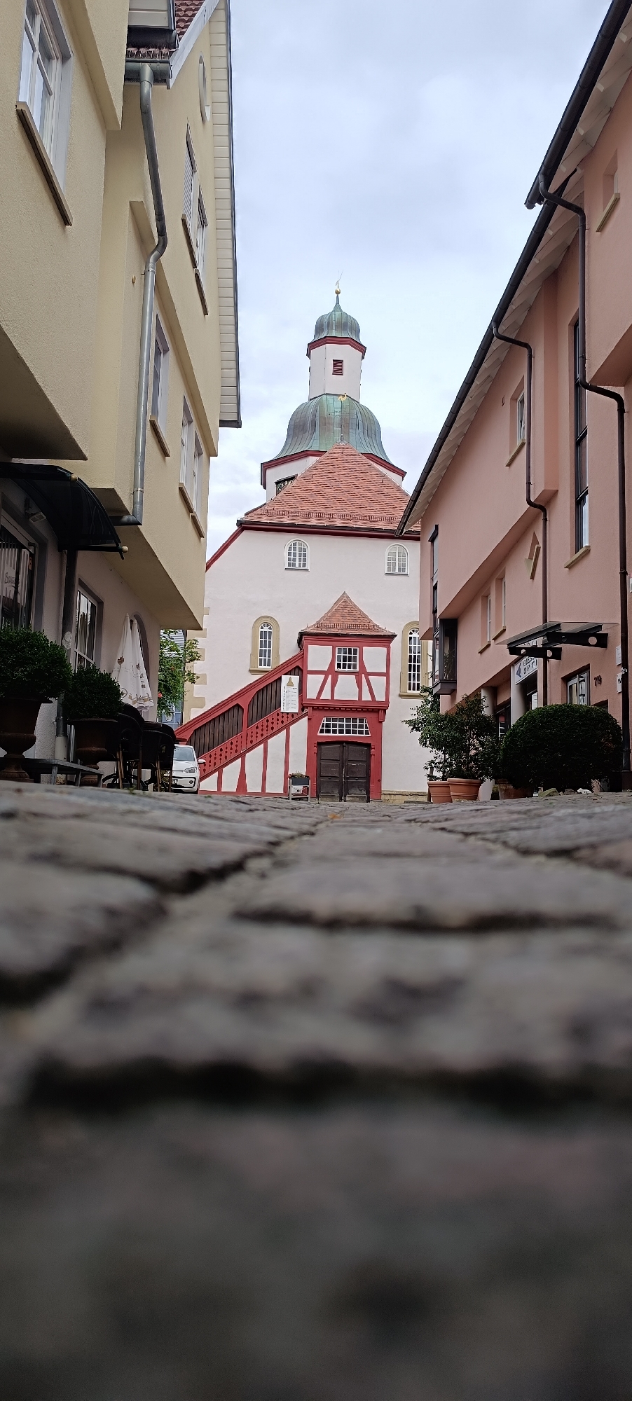This is a low-angle, eye-level shot of a cobblestone street in Winnenden, Germany, leading towards a white building with a prominent red-tiled roof and a green-domed tower. The building's facade features red and white half-timbering around the main entrance and a steep red staircase with white accents. The sky is overcast. On the left, a yellow building with windows and a gutter system is visible. On the right, a pink building with windows and awnings, including one with the signage "BEST OPTI", lines the street. Potted plants are situated in front of both the pink building and the white building's entrance area. A white car is parked in the background, behind the red staircase. The foreground is dominated by the texture and pattern of the cobblestones, which are slightly out of focus.