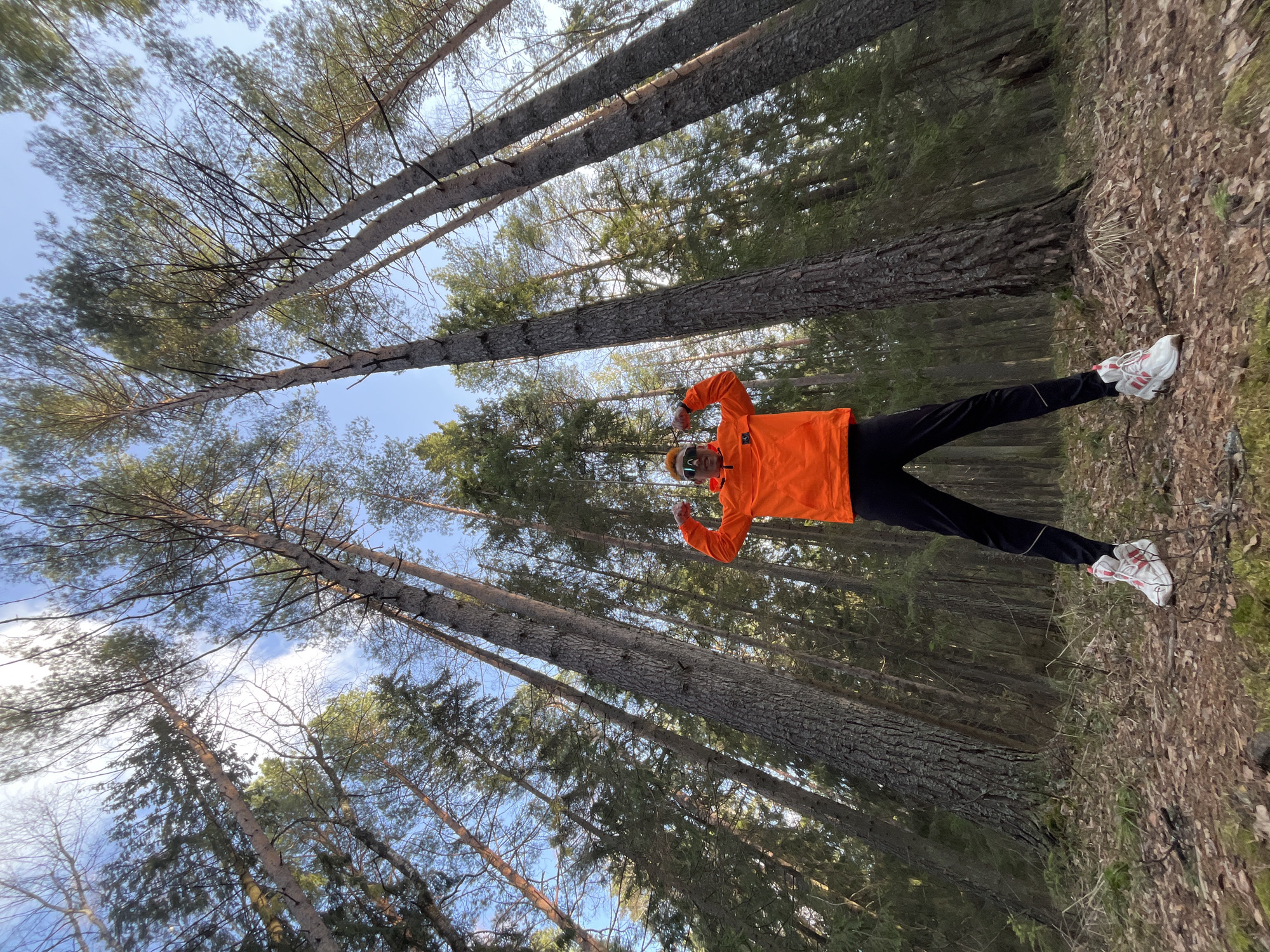 A person wearing an orange jacket, dark pants, white shoes, and reflective eyewear is standing in a forest. The ground is covered with leaves and the sky is visible between the trees. The trees are tall and appear to be in a coniferous forest, likely in the city of Chaikovsky, Russia, given the context. The person is standing with their arms raised and legs spread slightly apart, suggesting a celebratory or energetic pose.