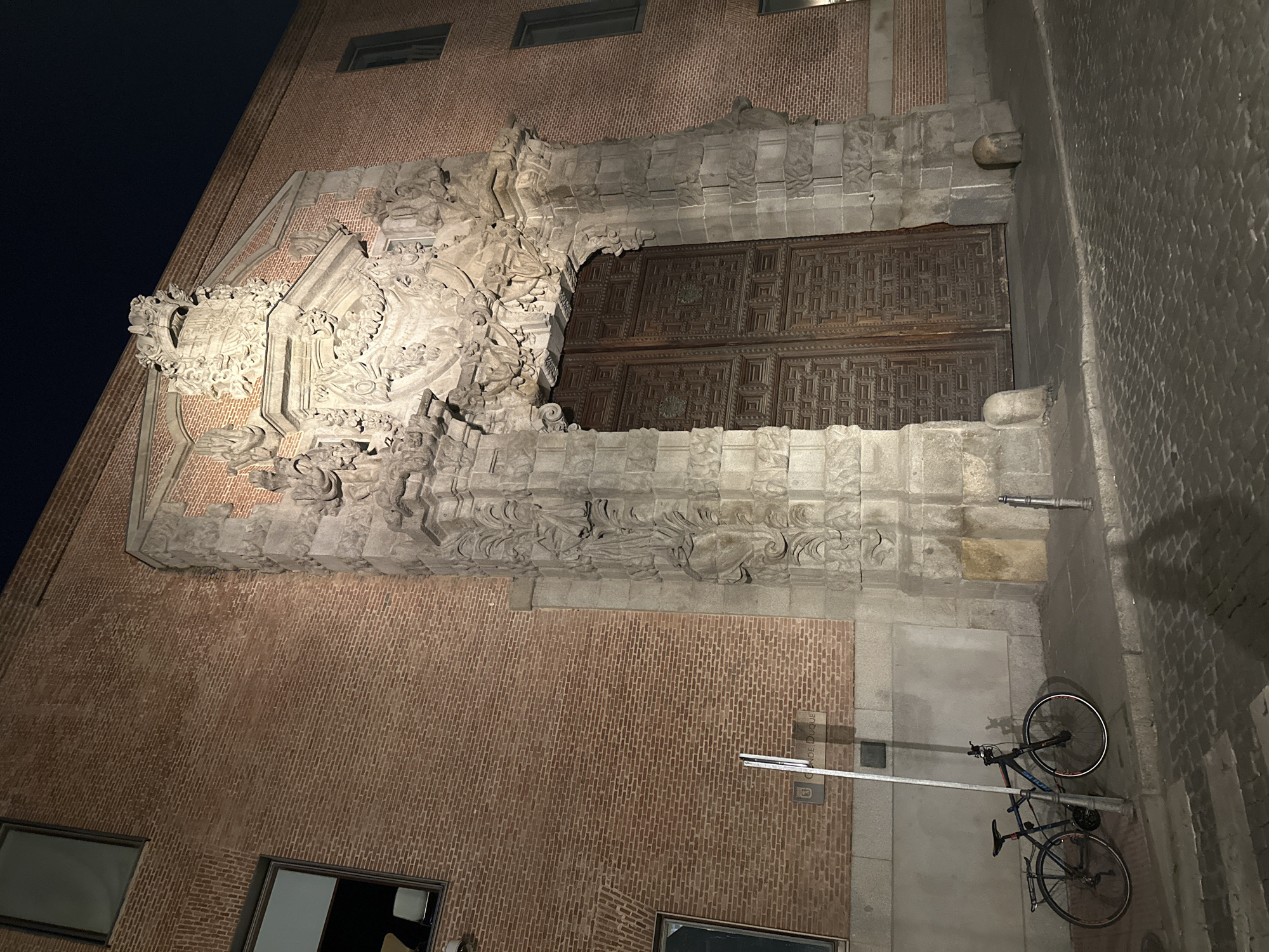 Exterior shot, slightly rotated, depicts an ornate building facade in Madrid, Spain, specifically the main entrance of the Conde Duque Cultural Center. The building is constructed of brick. The entranceway features a large, elaborately carved stone surround with a massive wooden door. Two bicycles are parked beside the building on the sidewalk. The sky is dark.