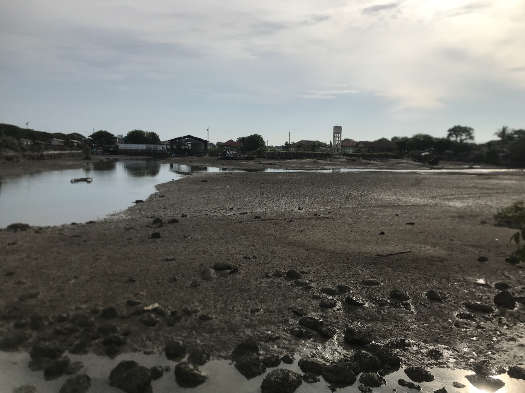 This image depicts a low-tide view of a coastal area in Lhokseumawe, Indonesia. The foreground is dominated by muddy, exposed tidal flats dotted with rocks and shallow pools of water reflecting the sky. In the middle ground, a body of water, likely part of the sea or a tidal channel, reflects a hazy sky with scattered clouds. The background reveals a small settlement with various structures, including low-rise buildings, some with corrugated metal roofs, and what appears to be a water tower. Vegetation, including trees and bushes, lines the edge of the settlement. The overall scene suggests a utilitarian or industrial coastal environment, possibly near a fishing village or a small port. There are no people visible in the image.